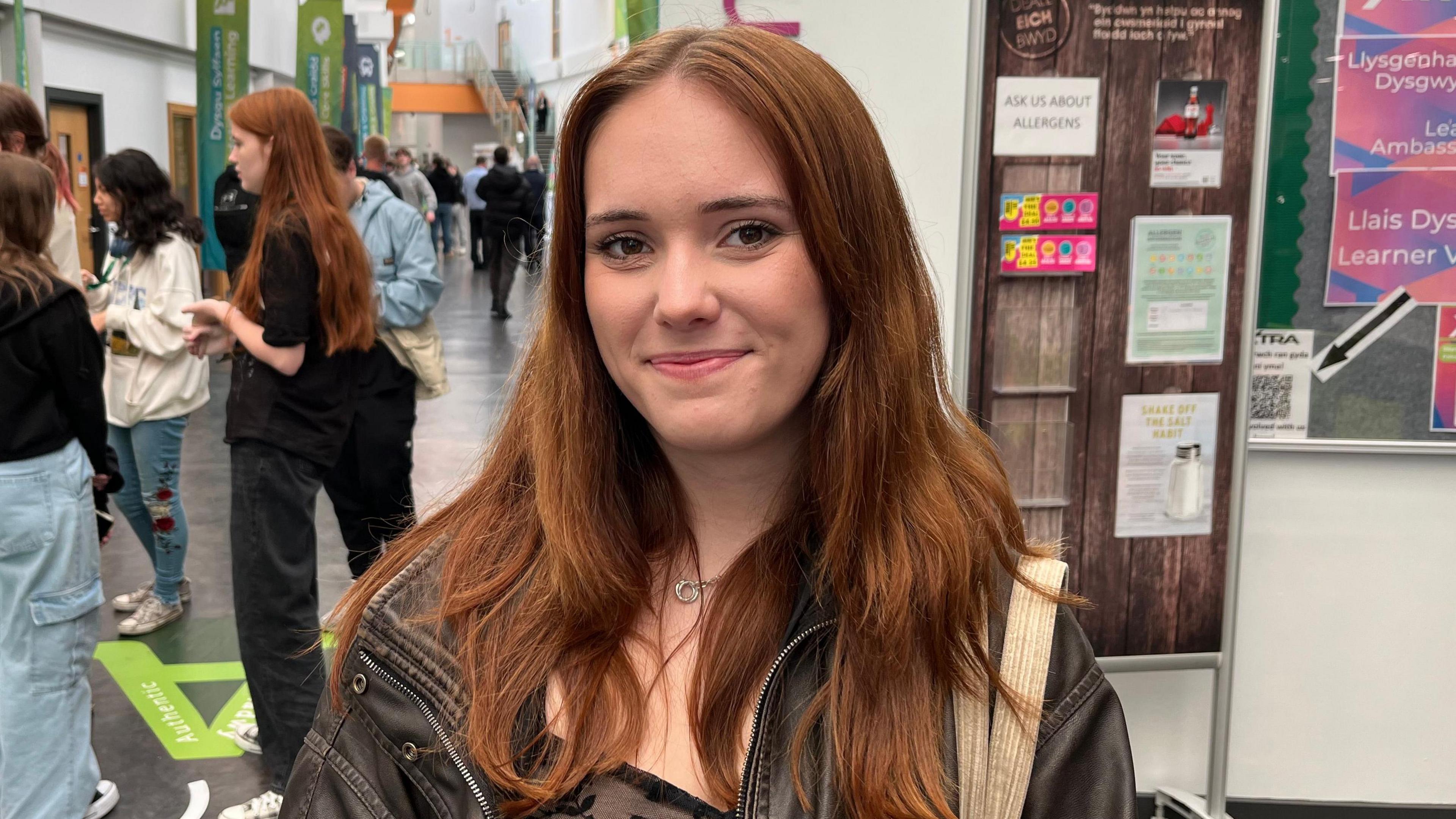Photograph of college student Joslin Parsons, 18, who has ginger mid length hair. She stands in a college hallway and smiles at the camera.