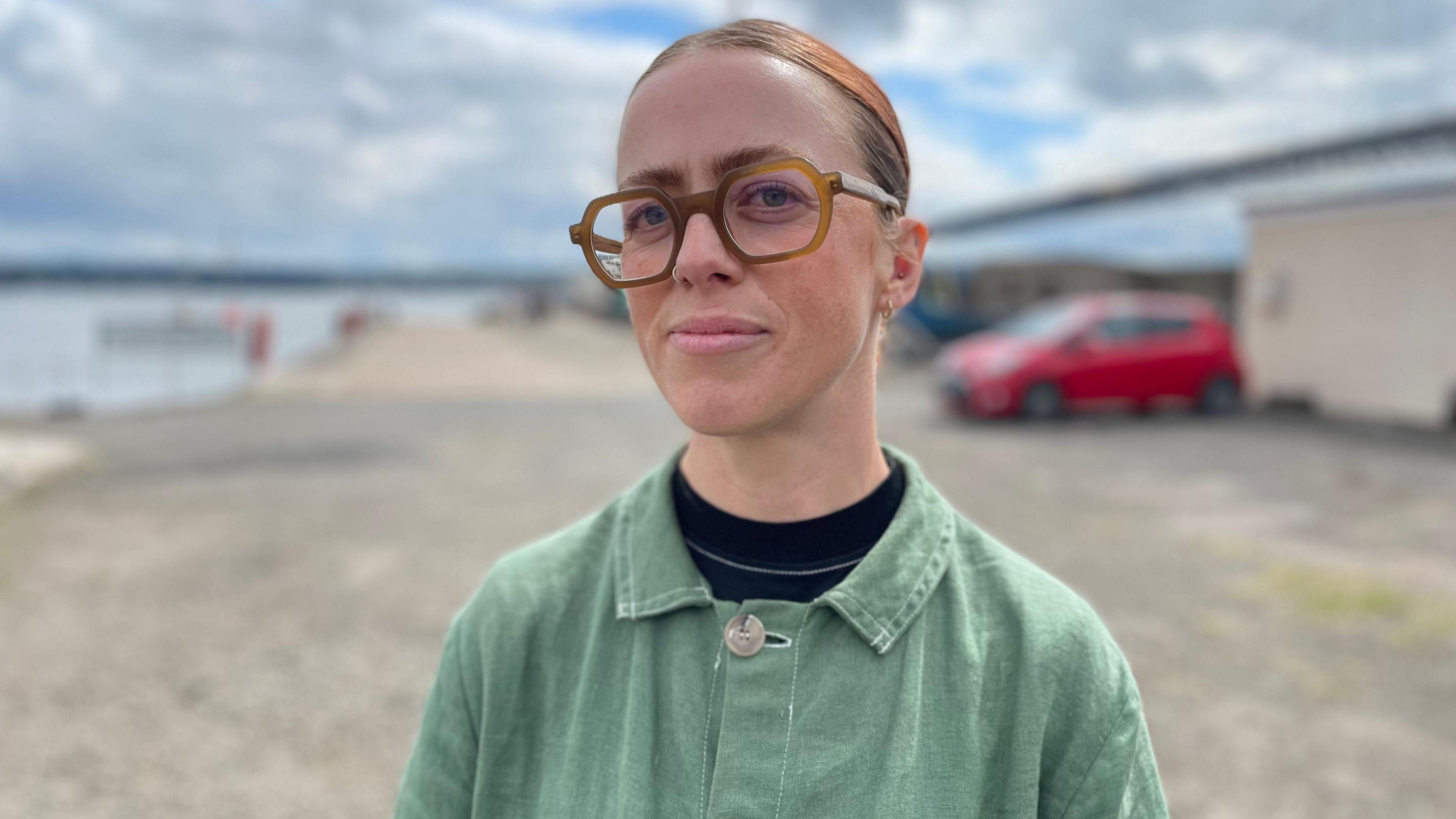 Amy Cameron looking straight at the camera. She has dark hair which has been tied back. She is wearing large orange coloured glasses on her face. She is pictured from the shoulders up and is wearing a light-green jacket over a dark top.