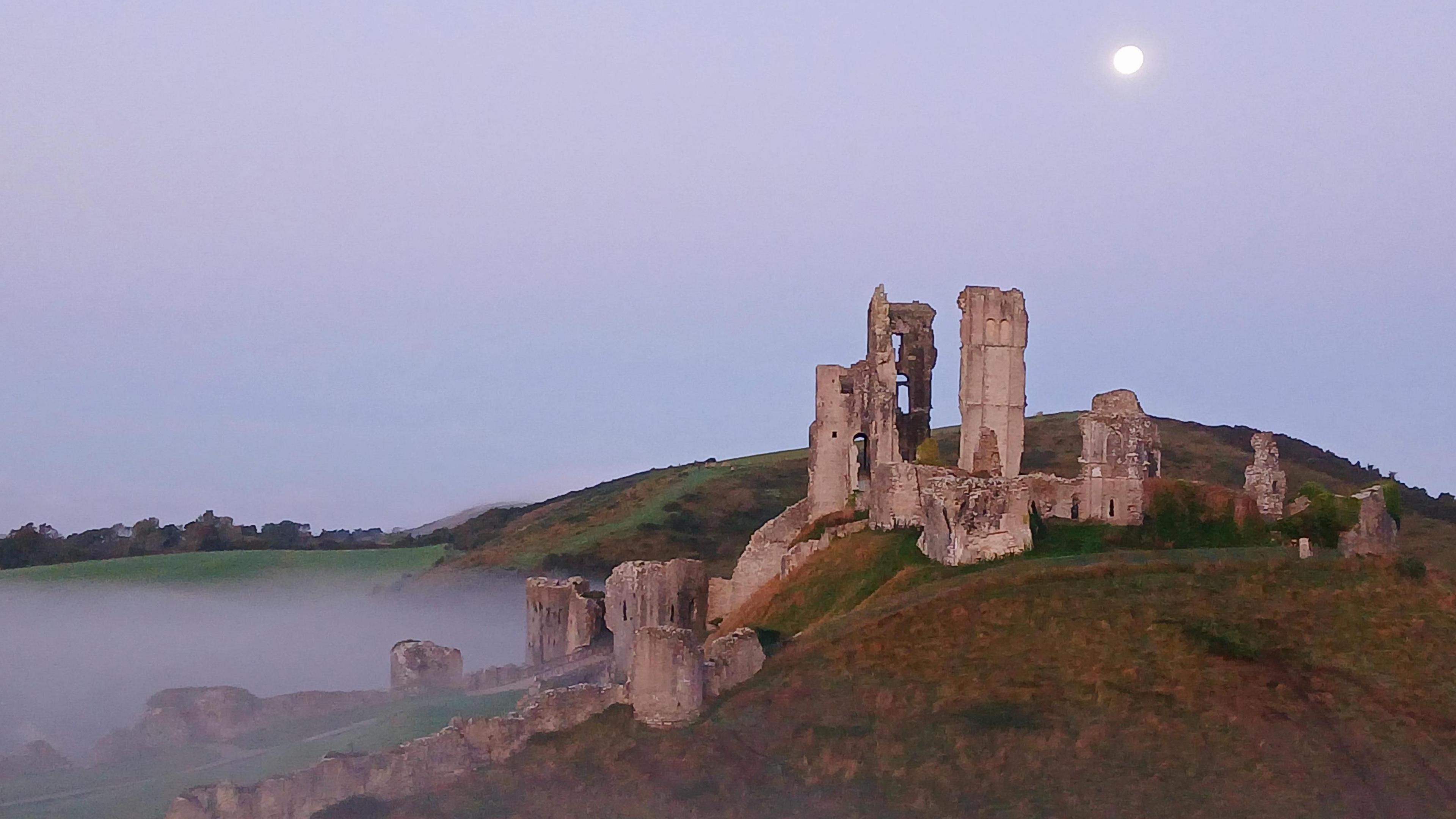 The wreck of Corfe Castle on top of a hill and surrounded by a mist and under a grey sky with the moon looming large in the sky above 