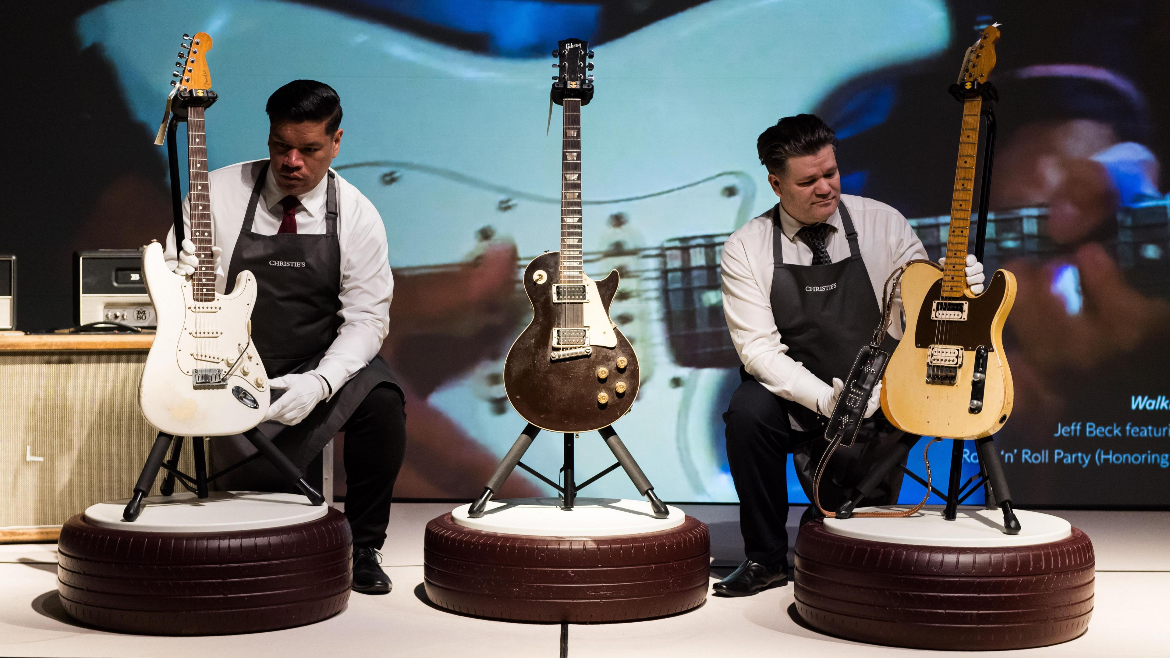 Two men in white shirts, white gloves, and black aprons, placing guitars on stands which are on top of fake car tyres. The guitar on the left is white, an already-place guitar in the middle is dark, and the guitar on the right is yellow.