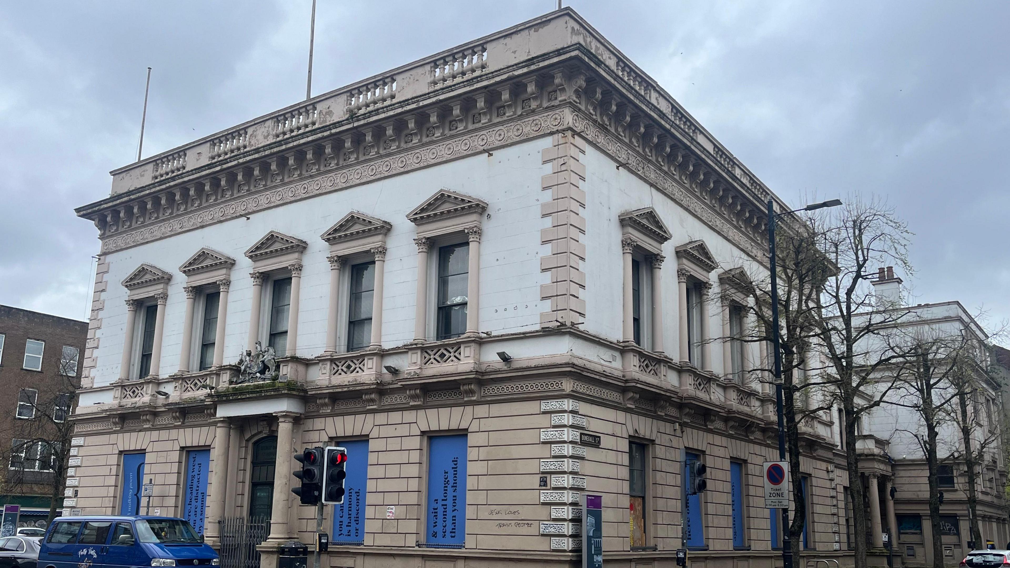 A beige and white square building with five windows on the first floor and a crest above the door. Each window has a pillar on either side, and there are a further four windows on the ground floor.