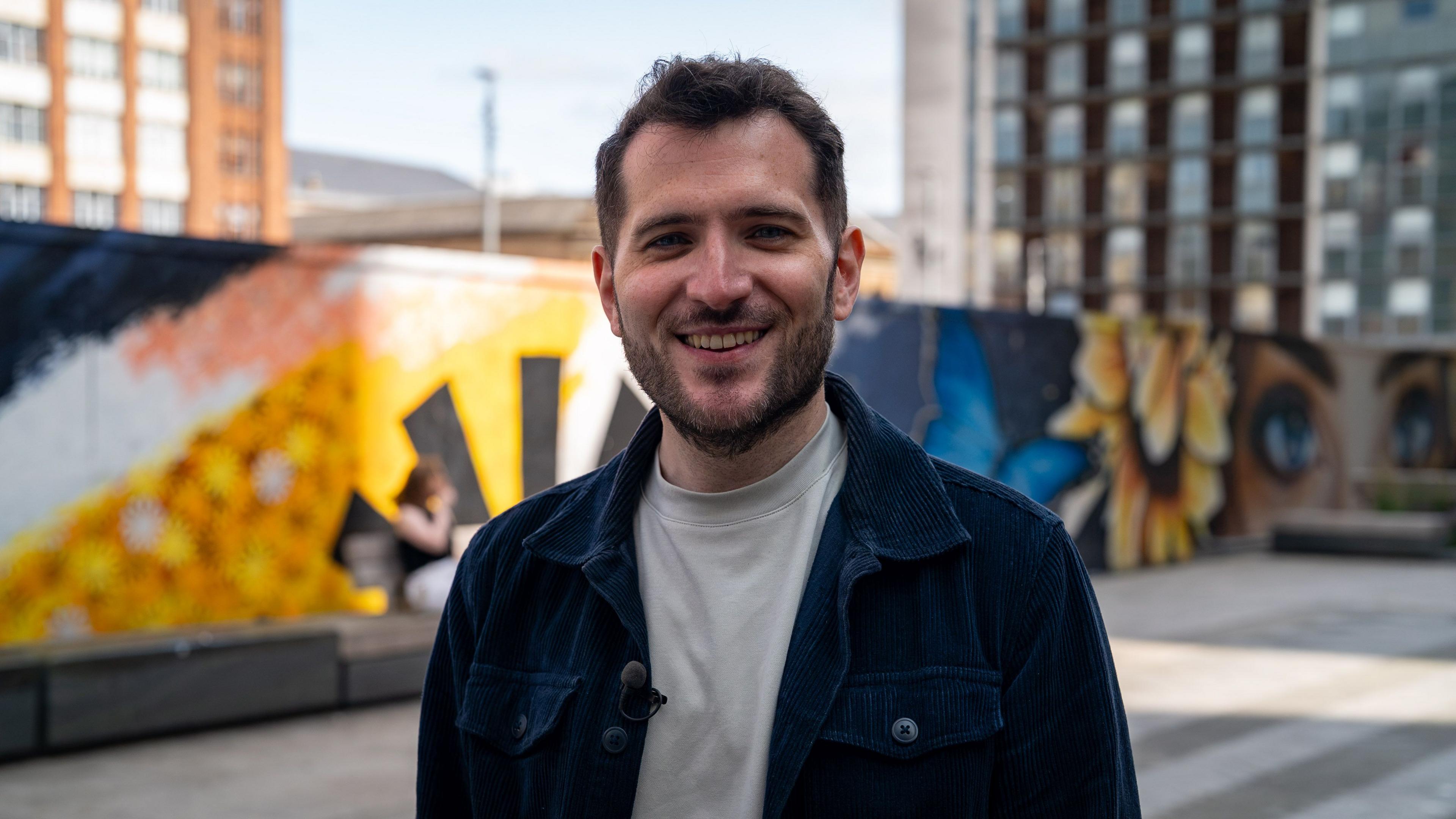Paul Sweeney standing in an urban plaza wearing a dark jacket and light-coloured top, with a colourful mural featuring flowers and butterflies and modern buildings in the background.
