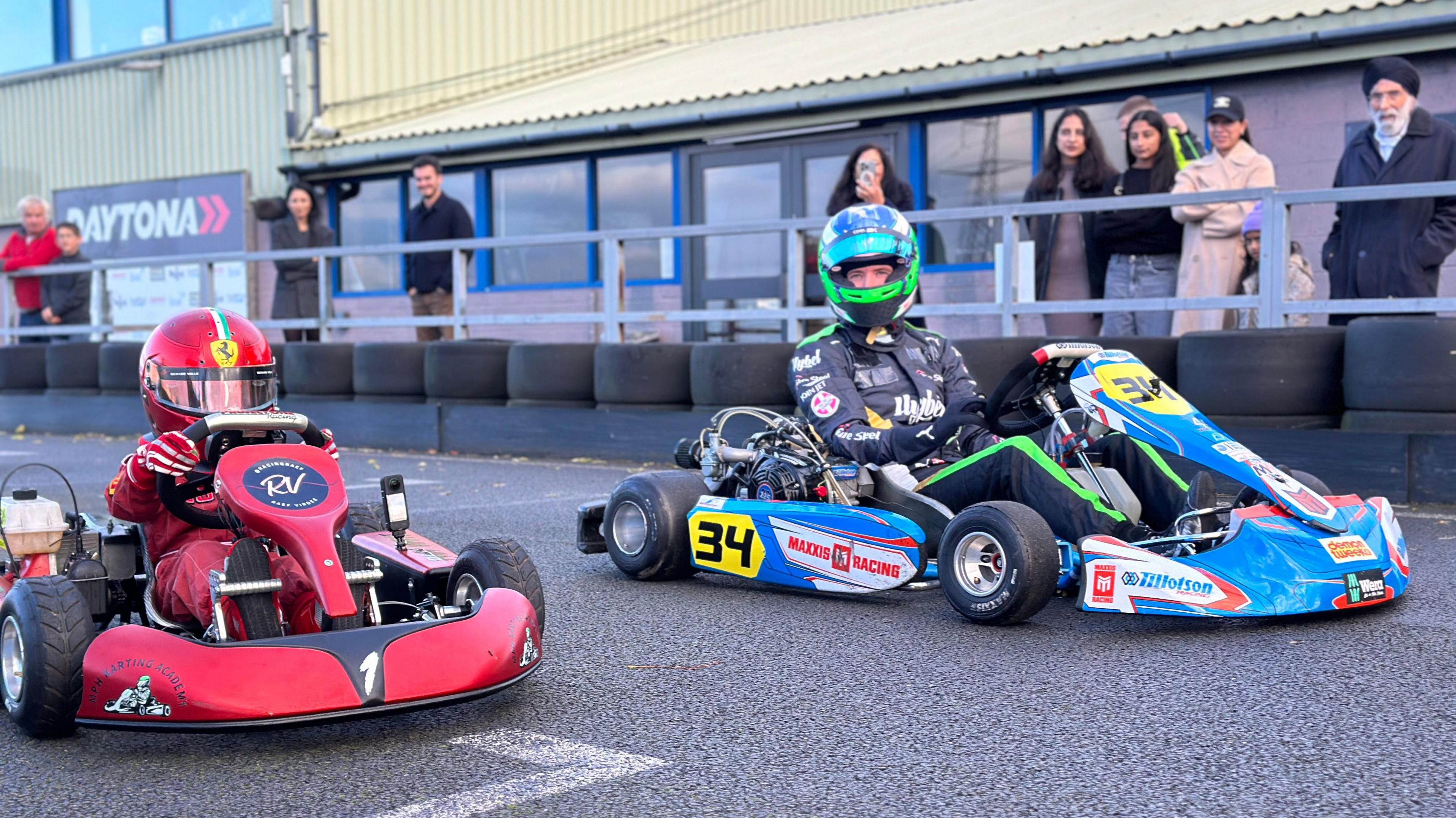 Raef is in his red racing suit and a red helmet, he is sitting on a red go kart. Vesti is next to him in a black and green racing suit, blue and green helmet, and is sitting in a blue kart. They are on the karting track with people spectating behind them