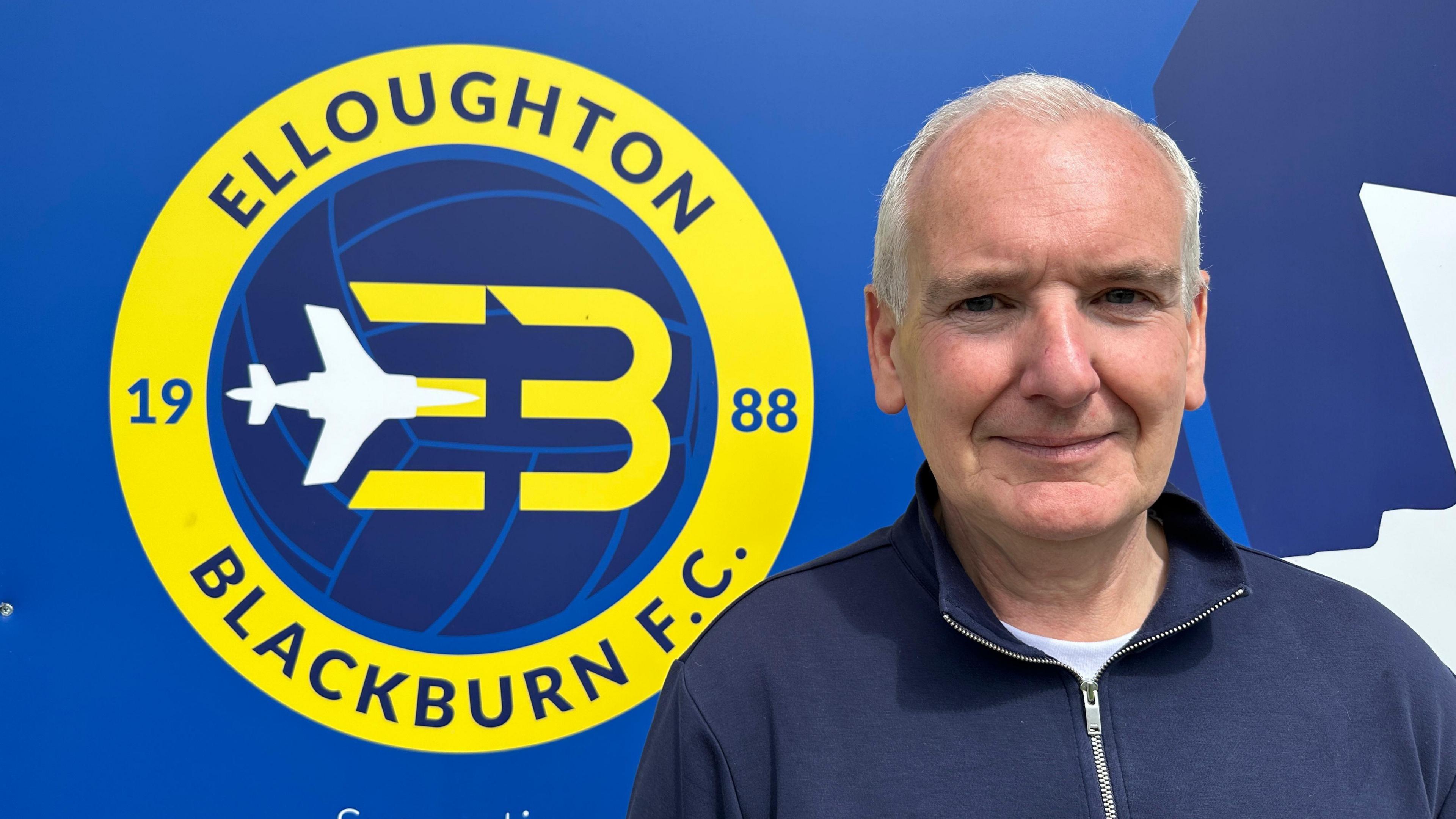Man with short white hair, a white t-shirt and blue top with zip. He is in front of a blue background and a yellow, white and blue Elloughton Blackburn F.C. 1988 logo.