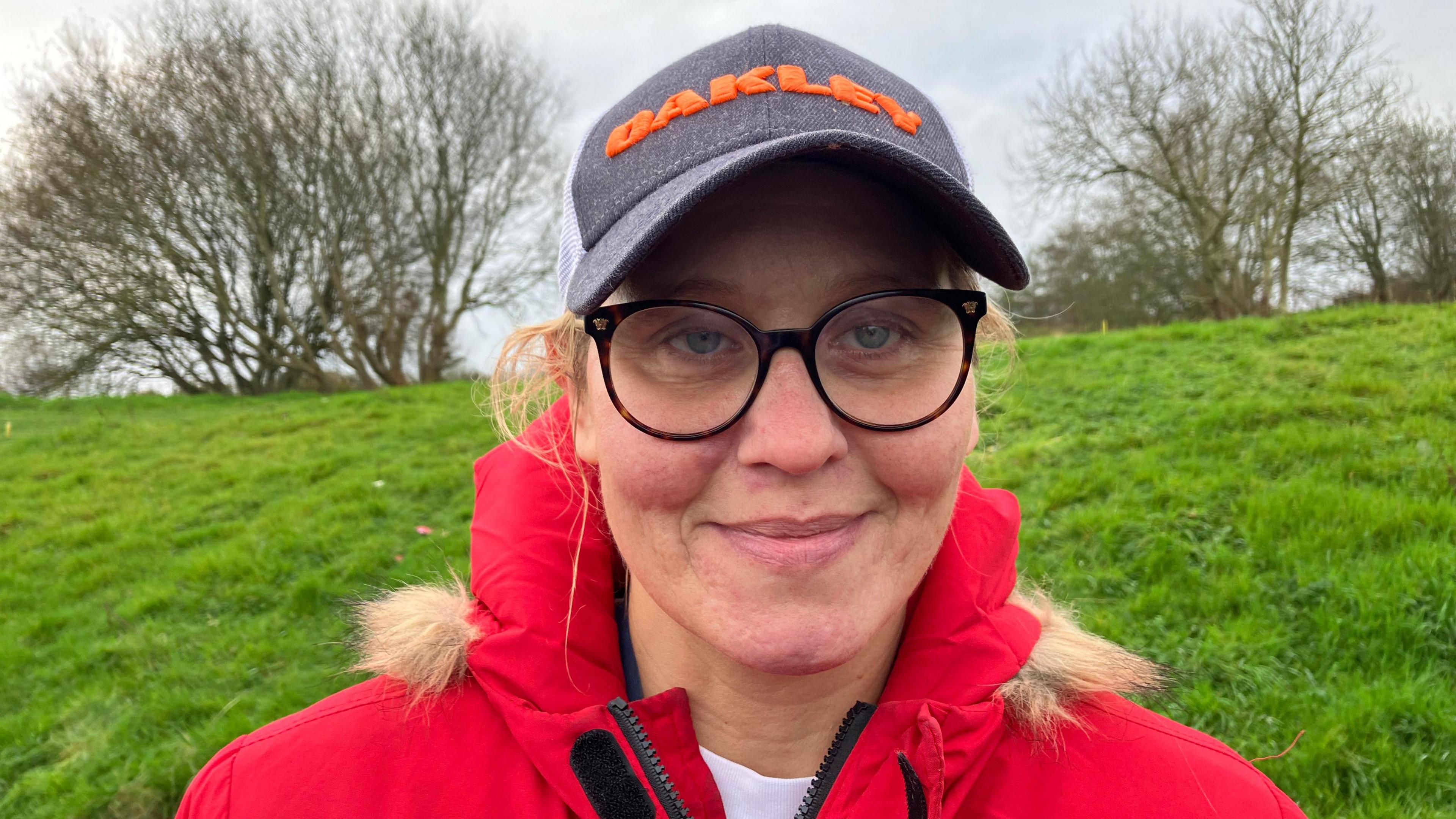 Nicki Sheller-Cullen pictured wearing a black cap and red coat. She is also wearing glasses and is standing at the bottom of a grassy slope. In the background is a number of trees.