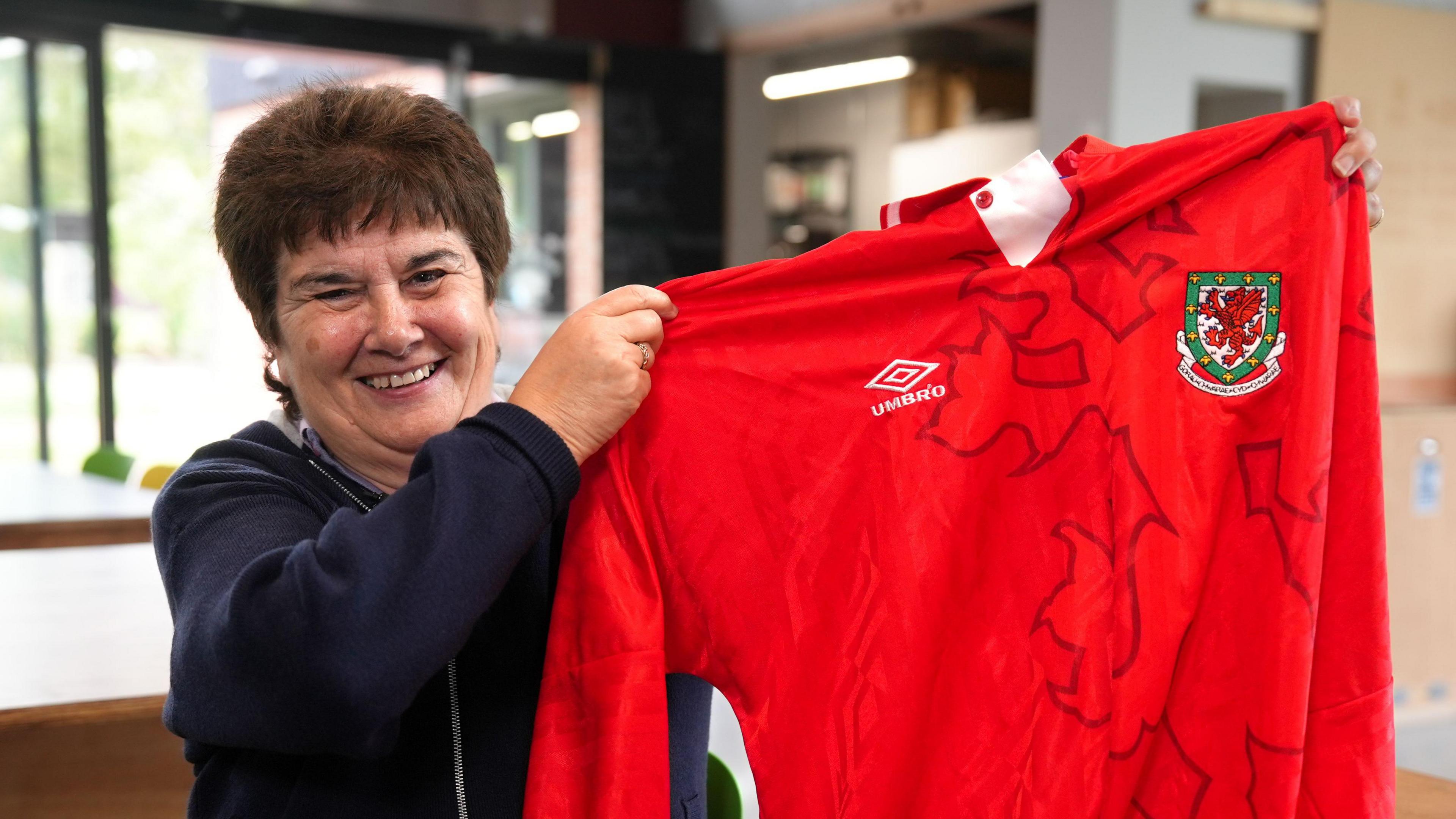 Michelle holds up her old red football jersey with long sleeves, an Umbro logo and a shield with the Welsh dragon on. She smiles at the camera widely. She has brown short hair.