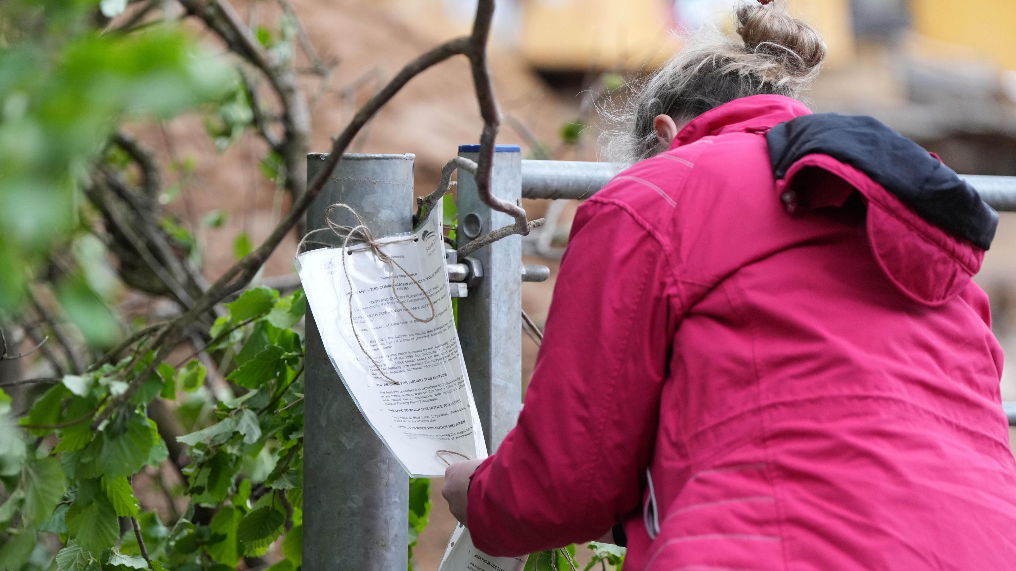 A woman wearing a pink coat. She is attaching a piece of paper to a metal gate with a piece of string.
