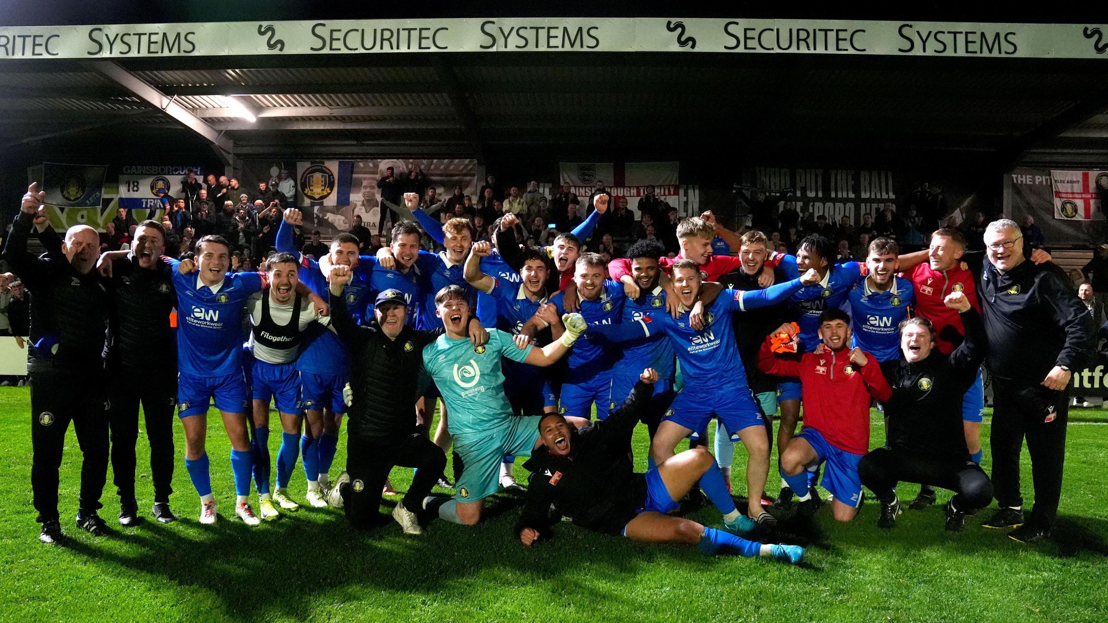 Football players in blue kits and coaches wearing black celebrate an FA Cup victory on the pitch in front of a stand.