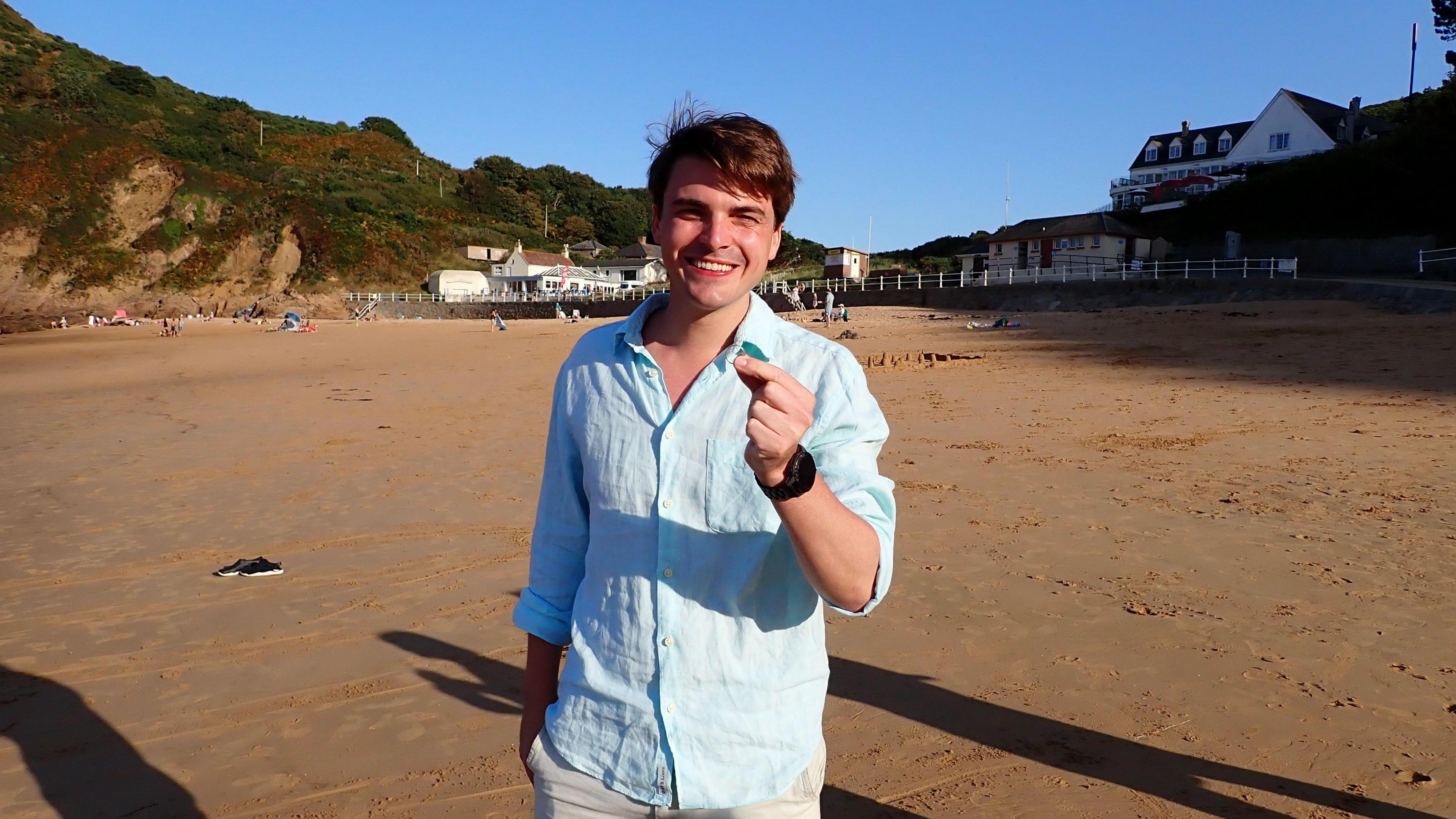 Ollie holding his wedding band in his fingers. He is wearing a blue shirt and is smiling at the camera. He is stood on a beach with the tide out. It is sandy and in the background are buildings.