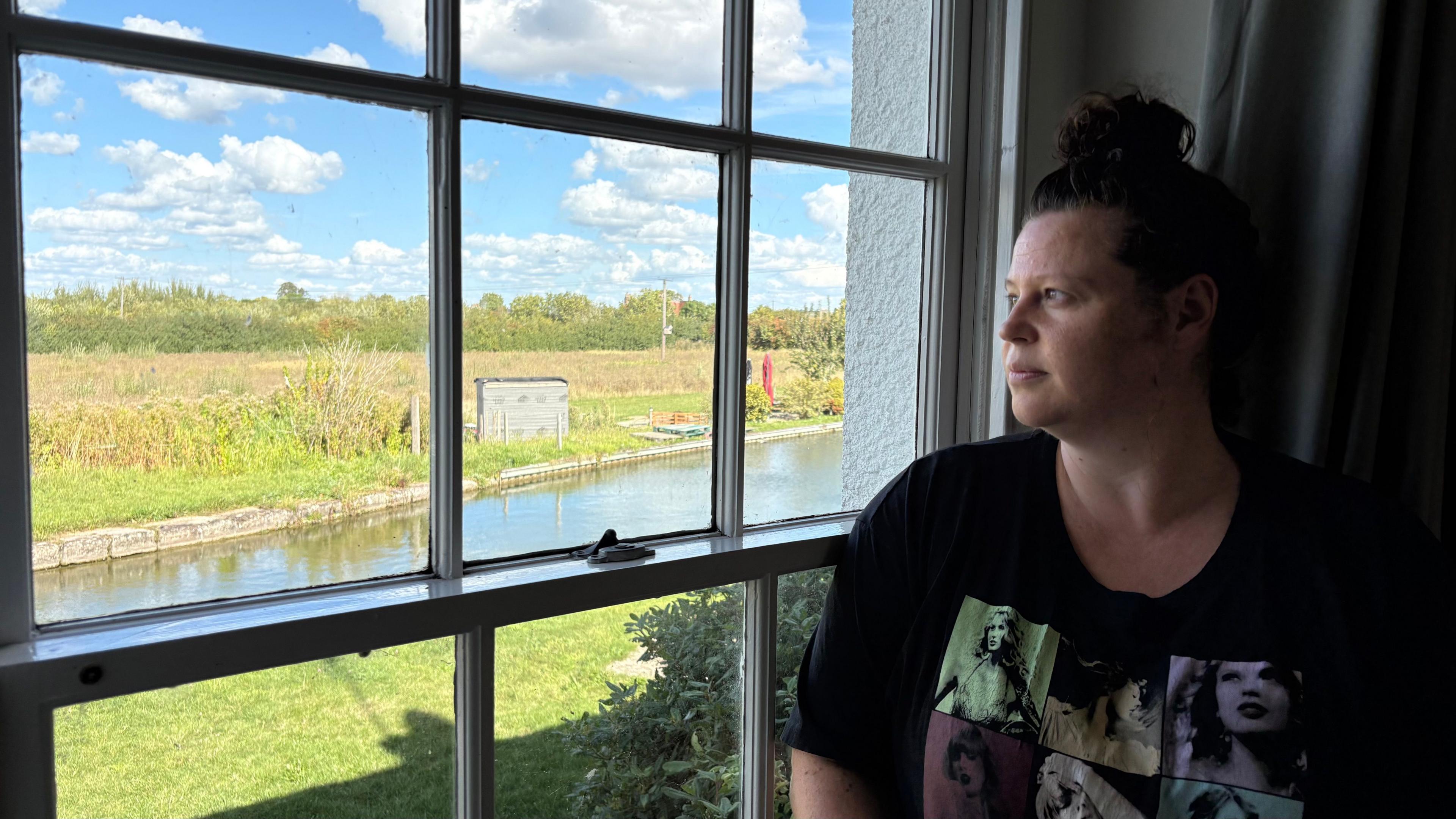 Kristina Hedley with long brown hair wearing a black T-shirt with green and blue graphics on the front. She is looking through a window made up of several square planes at a canal, with a grass and cereal field with a hedge behind.