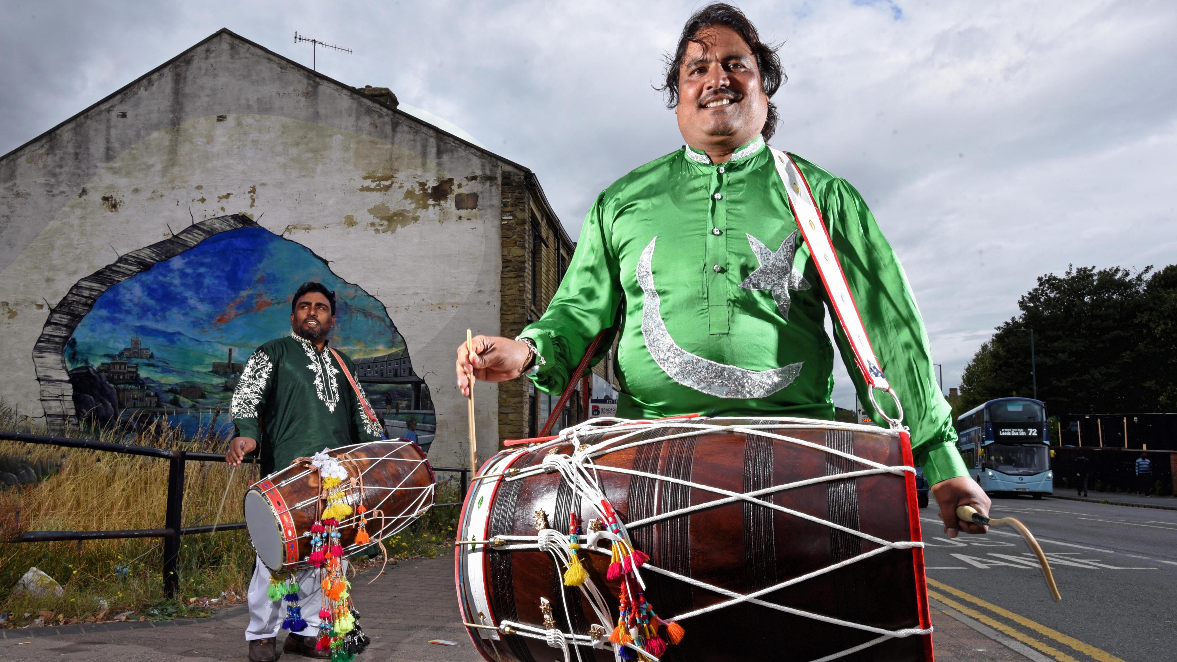 Two men dressed in traditional South Asian clothes and carrying dhol drums. Behind them is an urban streetscape, including a colourful mural.