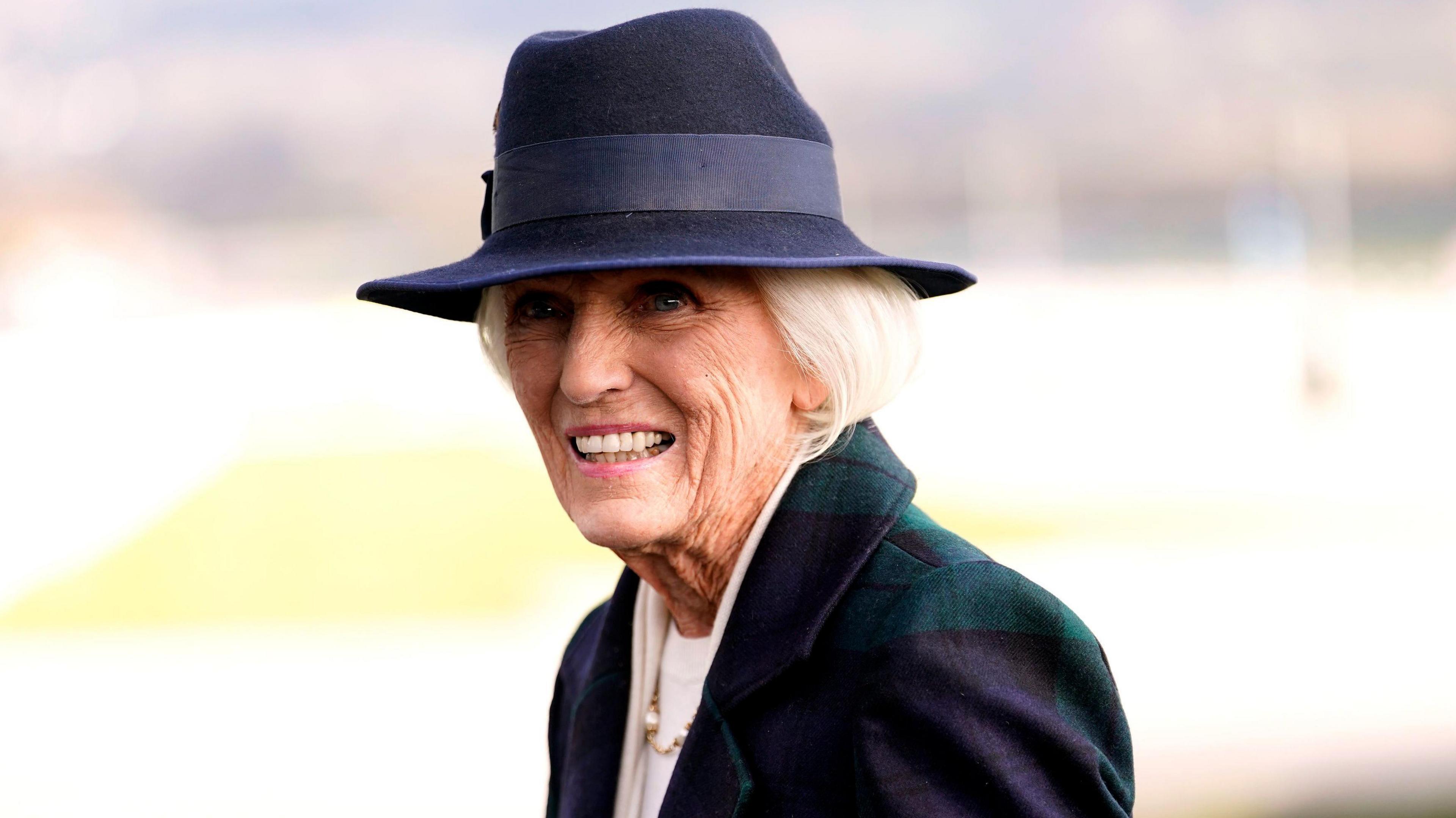 Mary Berry wearing a dark blue and green jacket, a dark blue sun hat and a white top. She has short white hair and smiles at the camera.