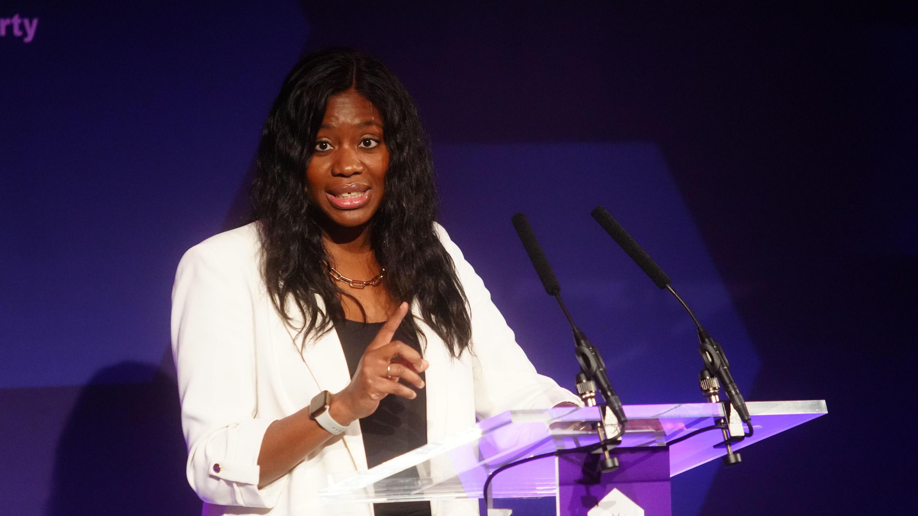 The minister for Devolution, Faith and Communities Miatta Fahnbulleh is wearing a white jacket and delivering a speech in front of a glass lectern with a purple backdrop.