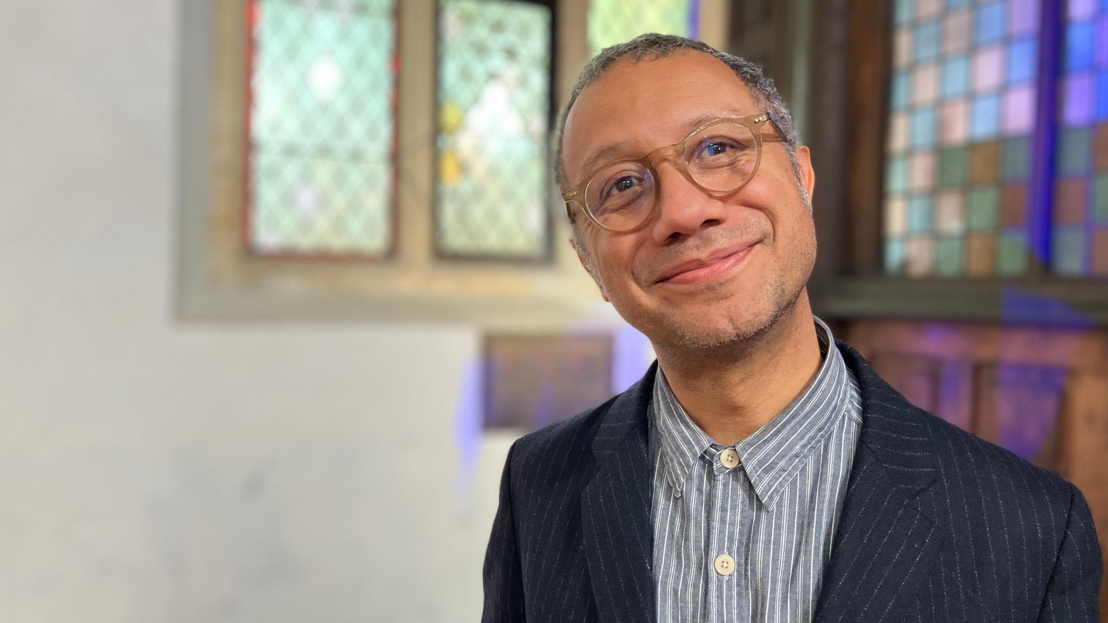 Robin Deacon stands smiling at the camera. He is inside a church with a stained glass window behind him. He is wearing clear glasses and a pinstripe jacket and buttoned-up shirt.