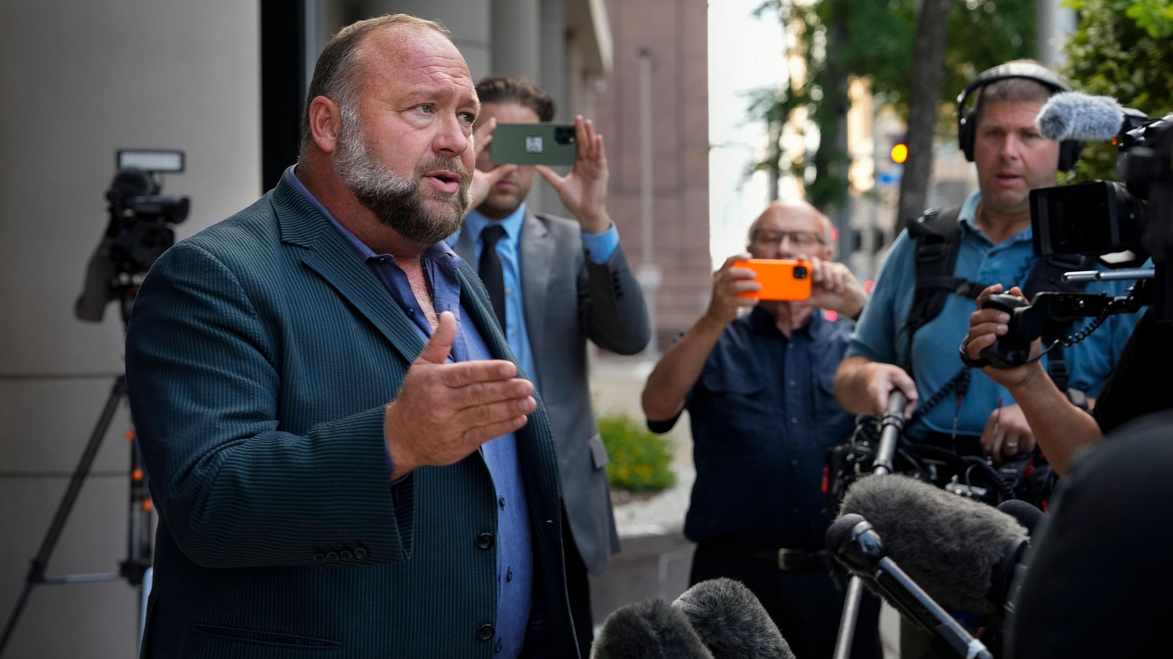 Alex Jones stood outside a court room in formal attire answering questions from the press. Microphones can be seen facing in his direction whilst someone takes a picture in the background.