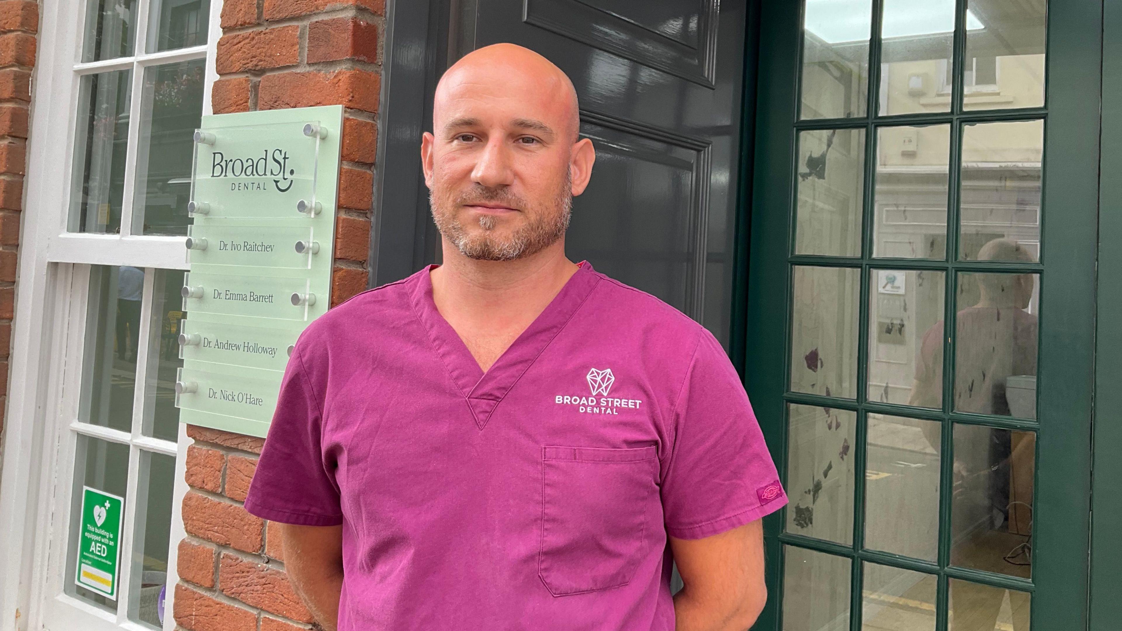 A man wearing a pink dentist shirt. He is looking at the camera and has his arms behind his back. He is stood in front of his practice which has a dark green door and a white window. There is a off-white sign to his left with the words 'Broad St. Dental'.