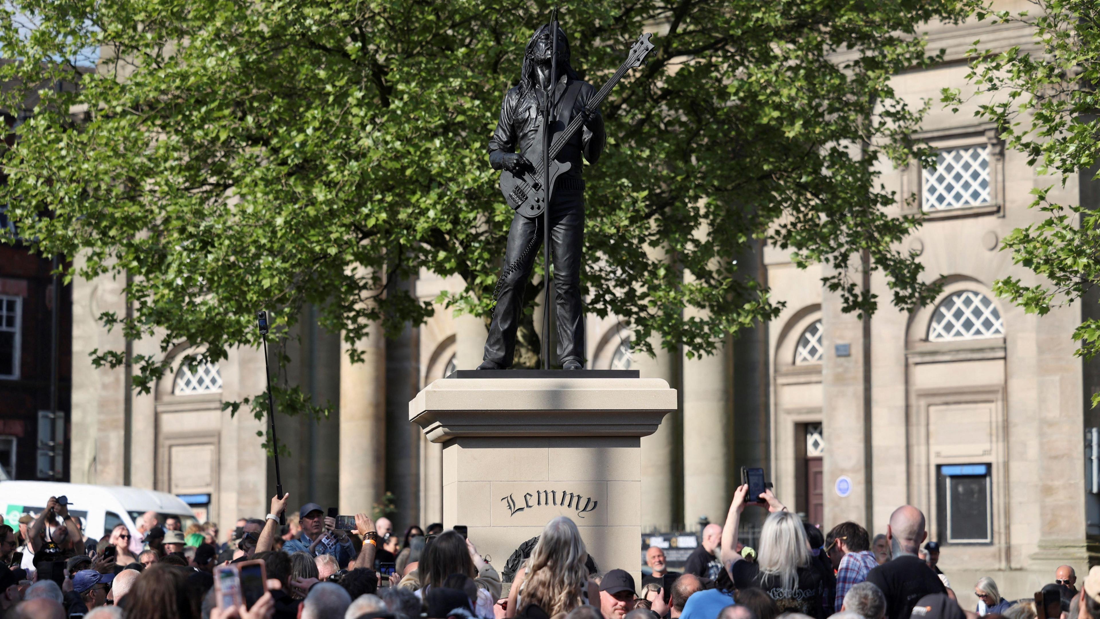 A crowd, some of them holding up phones, gathers around a beige-coloured plinth which has a black statue on top of Lemmy - a man with long hair holding a bass guitar and singing upwards into a microphone. A large building can be seen in the background.
