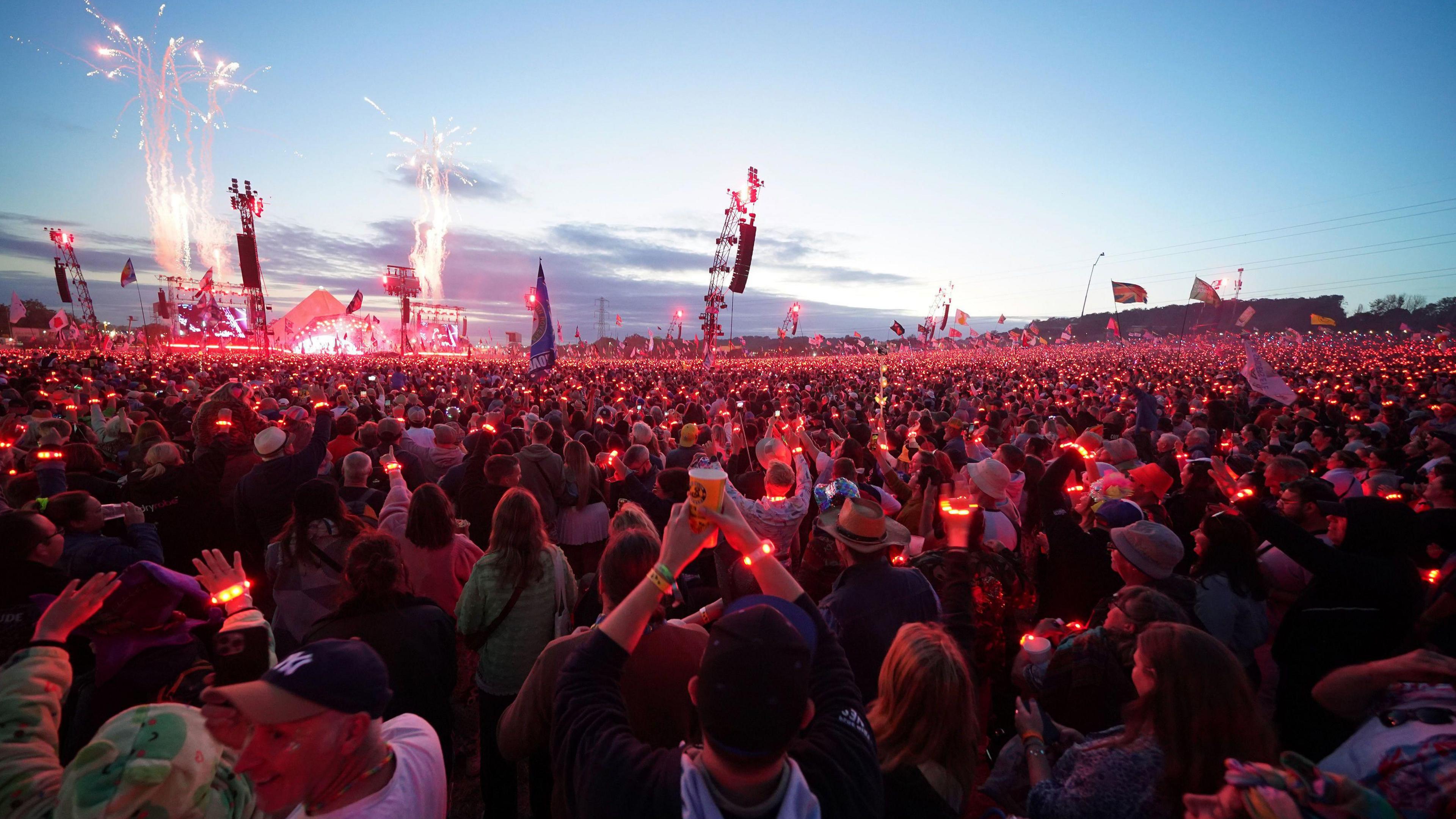 A crowd of thousands of people enjoy watching Coldplay on the Pyramid Stage at Glastonbury wearing red light-up wristbands,