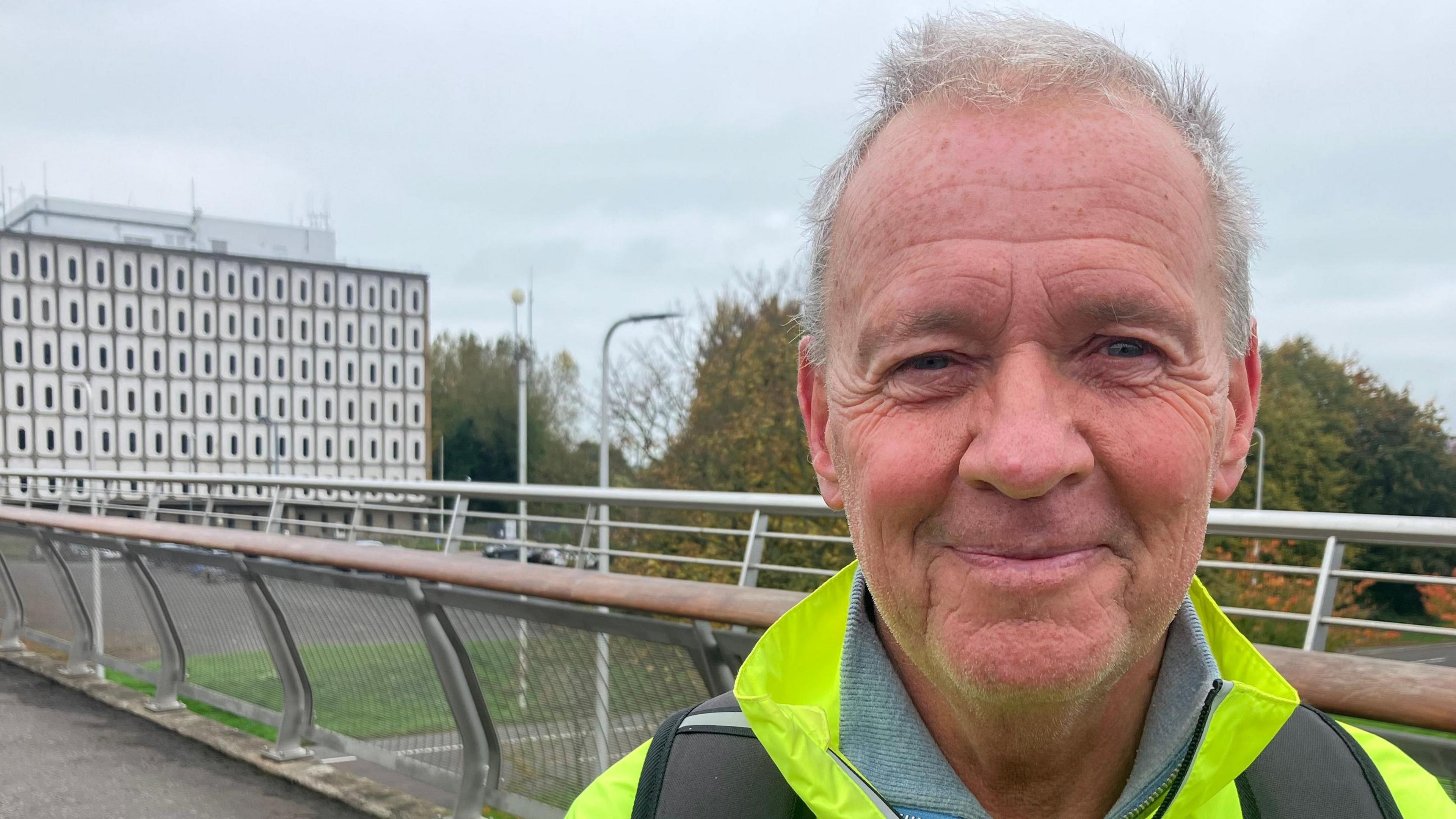 Ronnie Hanna pictured at a bridge leading to Marlborough House. He is wearing a hi-vis jacket and a backpack. He smiles at the camera and has short white hair.