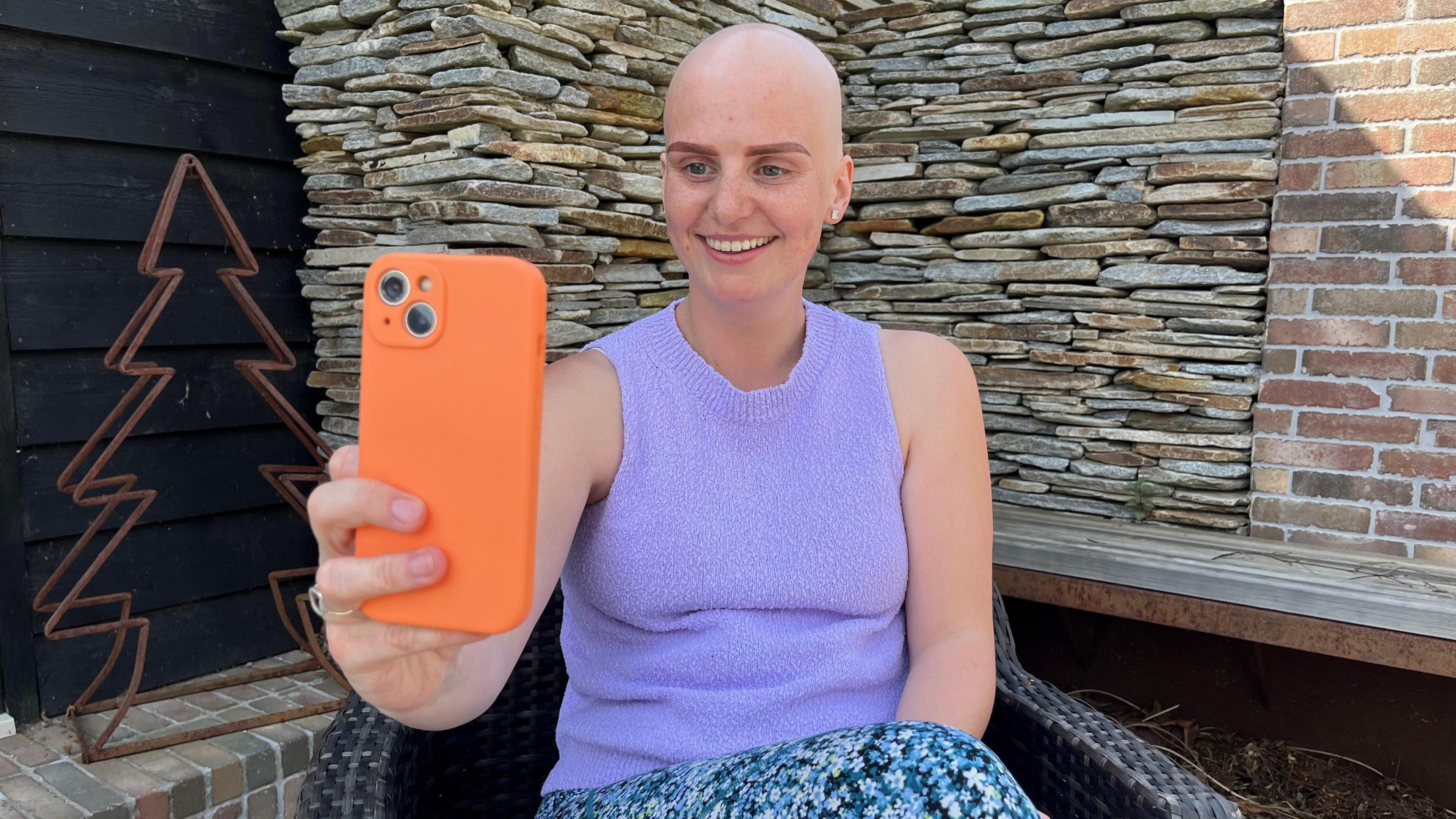 A young girl dressed in a purple top and multi-coloured trousers smiles into an iPhone with a bright orange case.