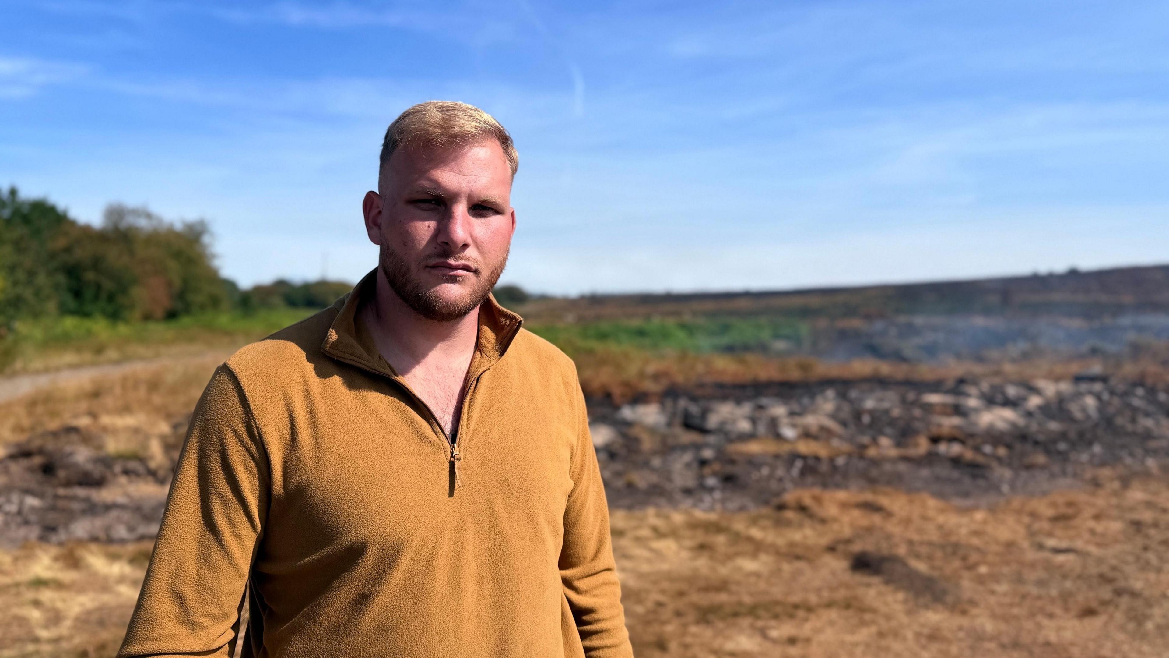 A man with blond hair stood in front of a burnt field. He's wearing a mustard yellow quarter-zip. The sky above is bright blue. 