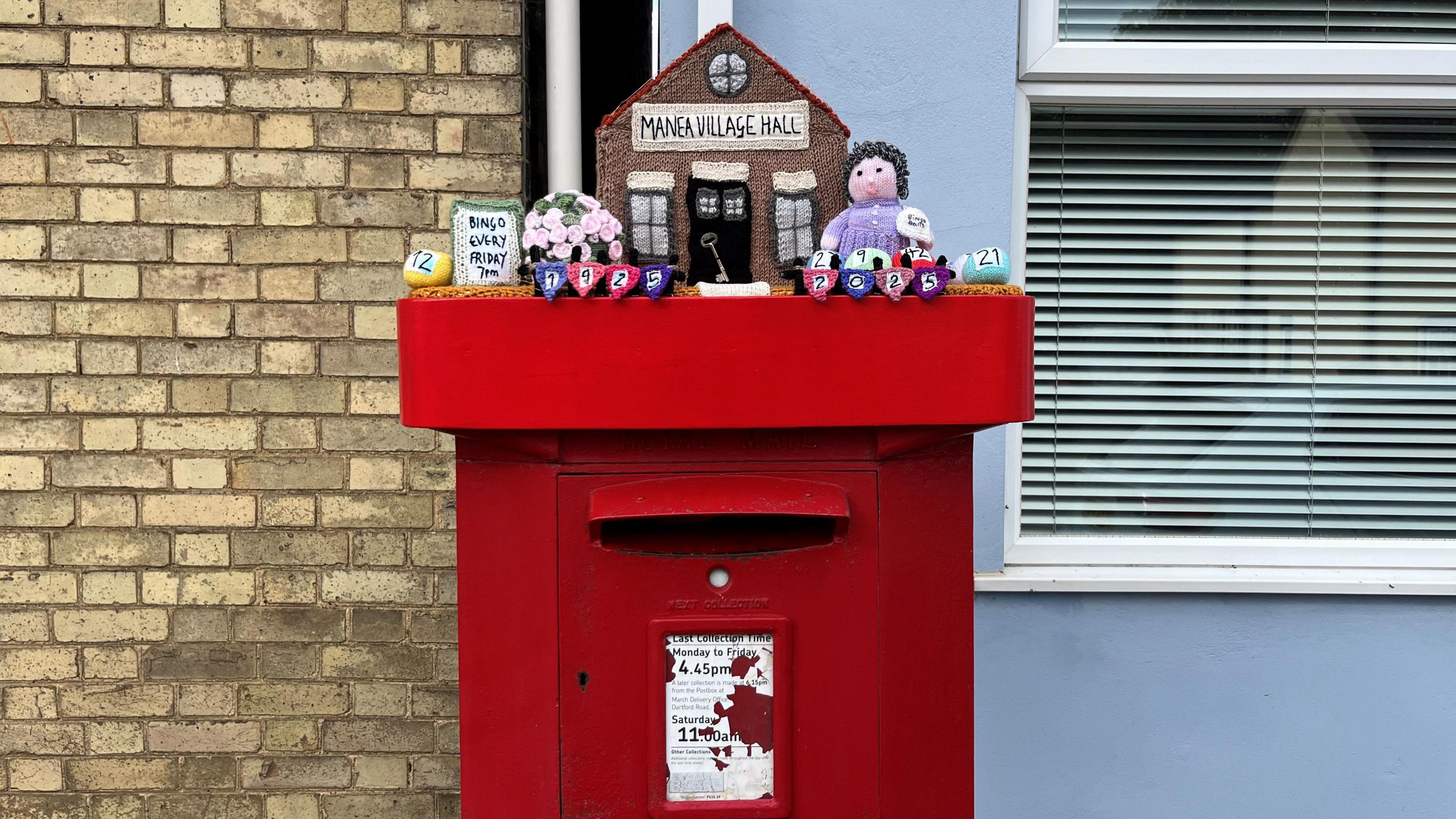 A colourful woollen topper on a square red post box with housing behind it. There is a model made of wool of the Manea village hall with various woollen items around it.