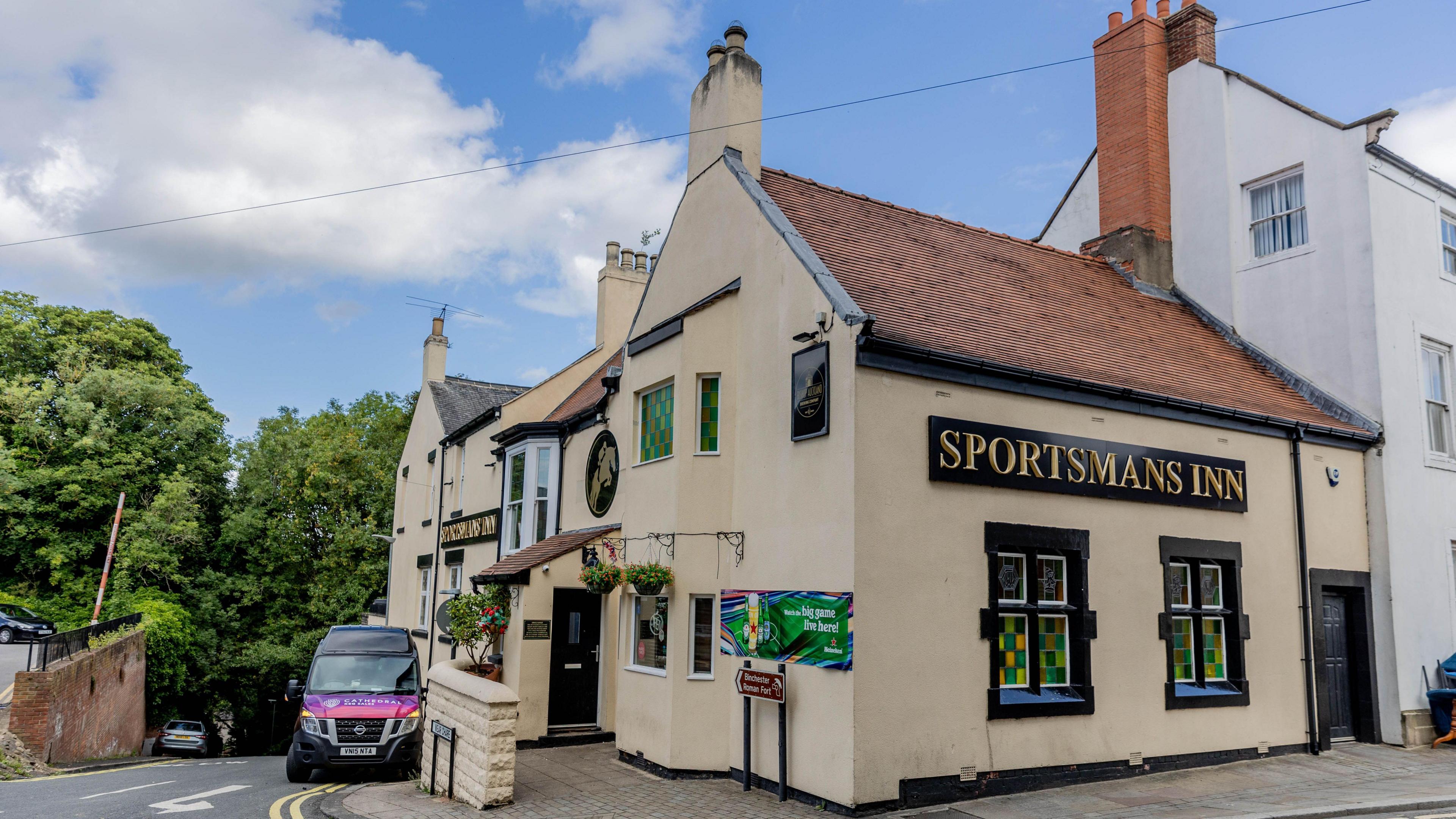 Large yellow pub building, which looks like a house, with a tiled slanted roof. There is a large black sign at the front of the building which says Sportsmans Inn in gold writing. There is a van parked outside the pub.