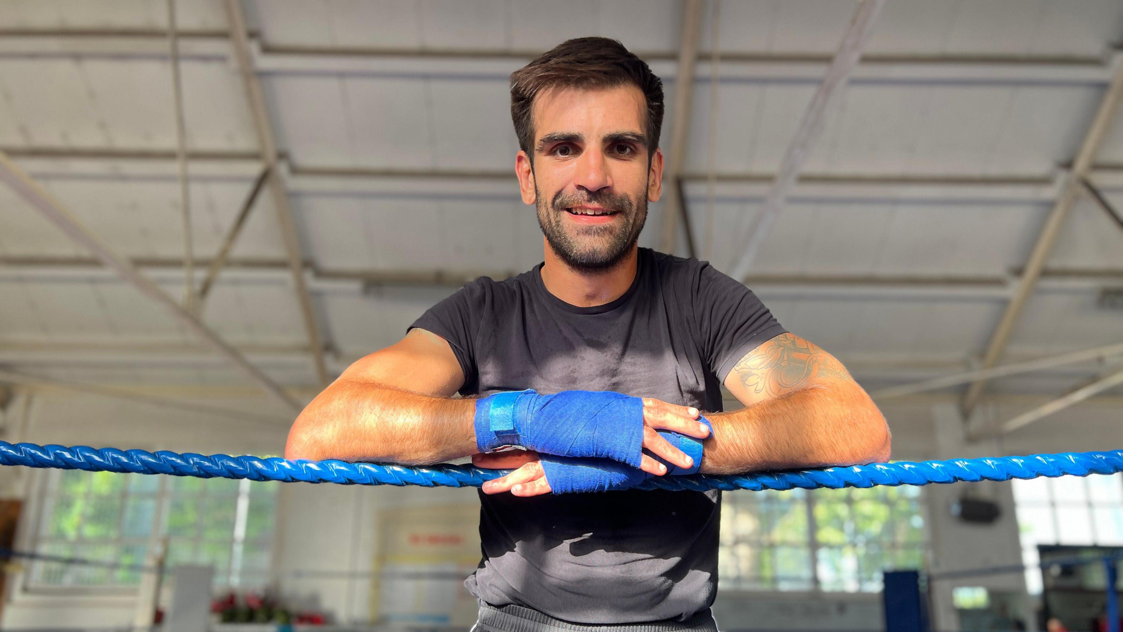 Chris leans on the ropes in a boxing ring as he looks down and smiles at the camera. He has brown hair and a beard and is wearing a black t-shirt and his hands have blue tape around them.