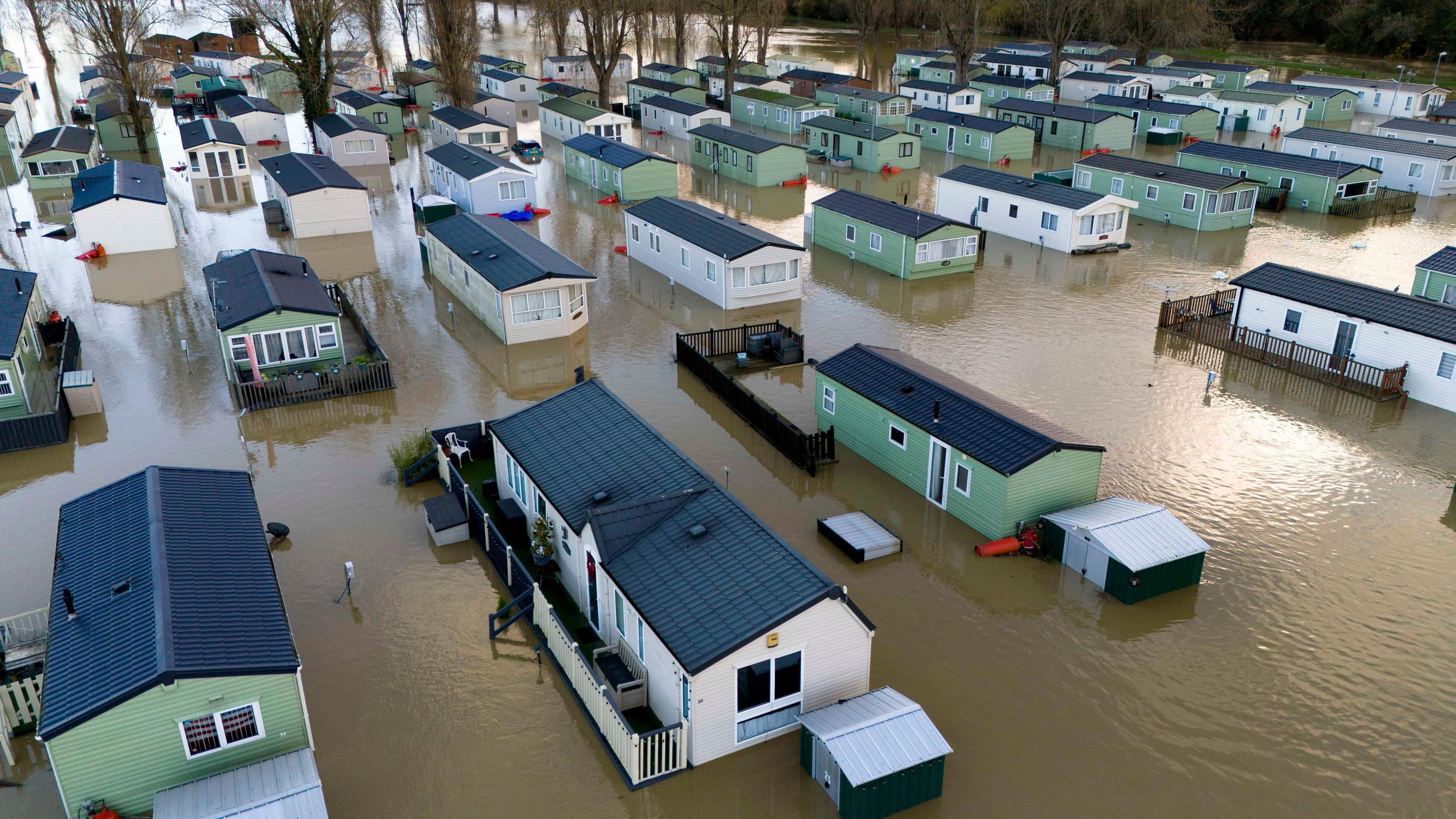 Green and white static caravans, which are dotted between trees, are seen deluged by brown floodwater which is at door height