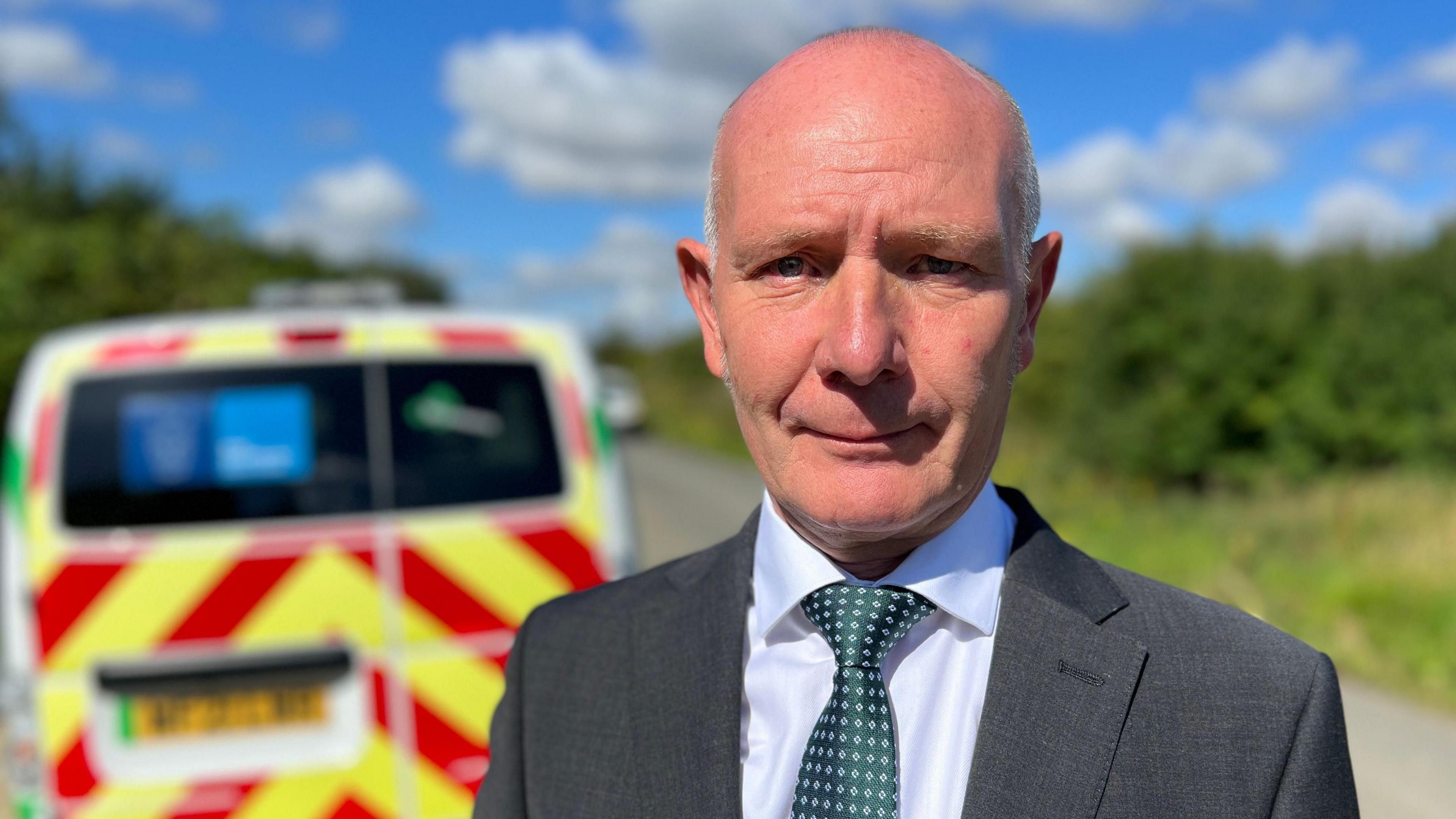 Darryl Preston standing outside with a police van behind him. He has cropped white h air and is wearing a grey suit, white shirt and green tie