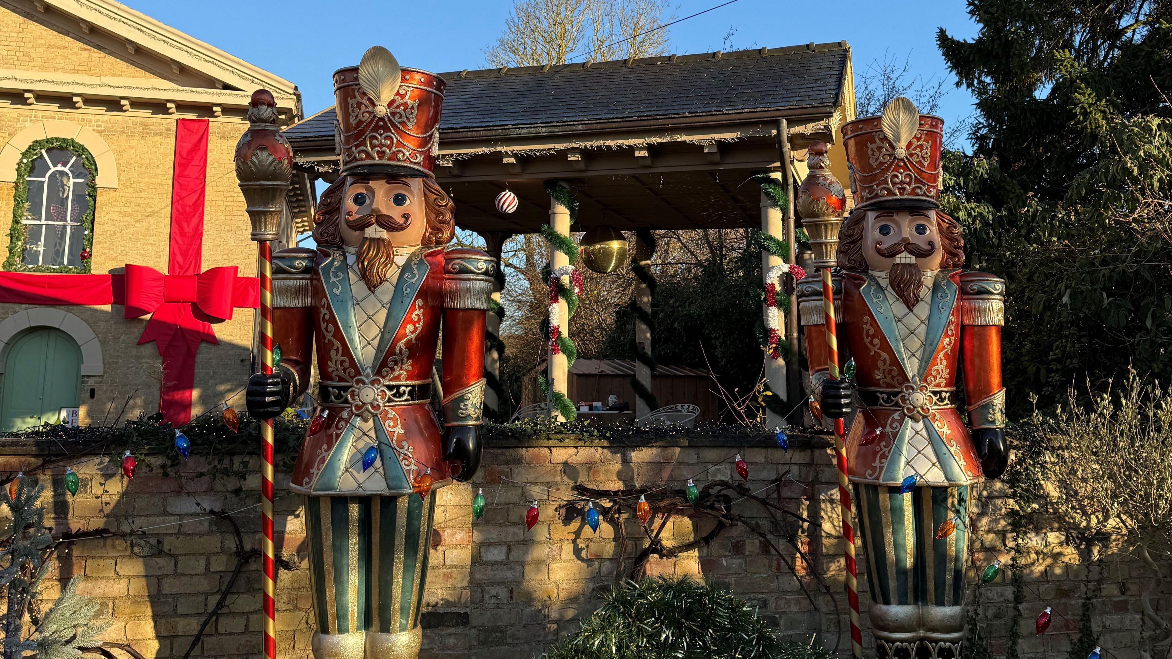 Two Christmas nutcracker soldier models are standing in front of a house. There is a wall behind them and a stone well with Christmas decorations on it.