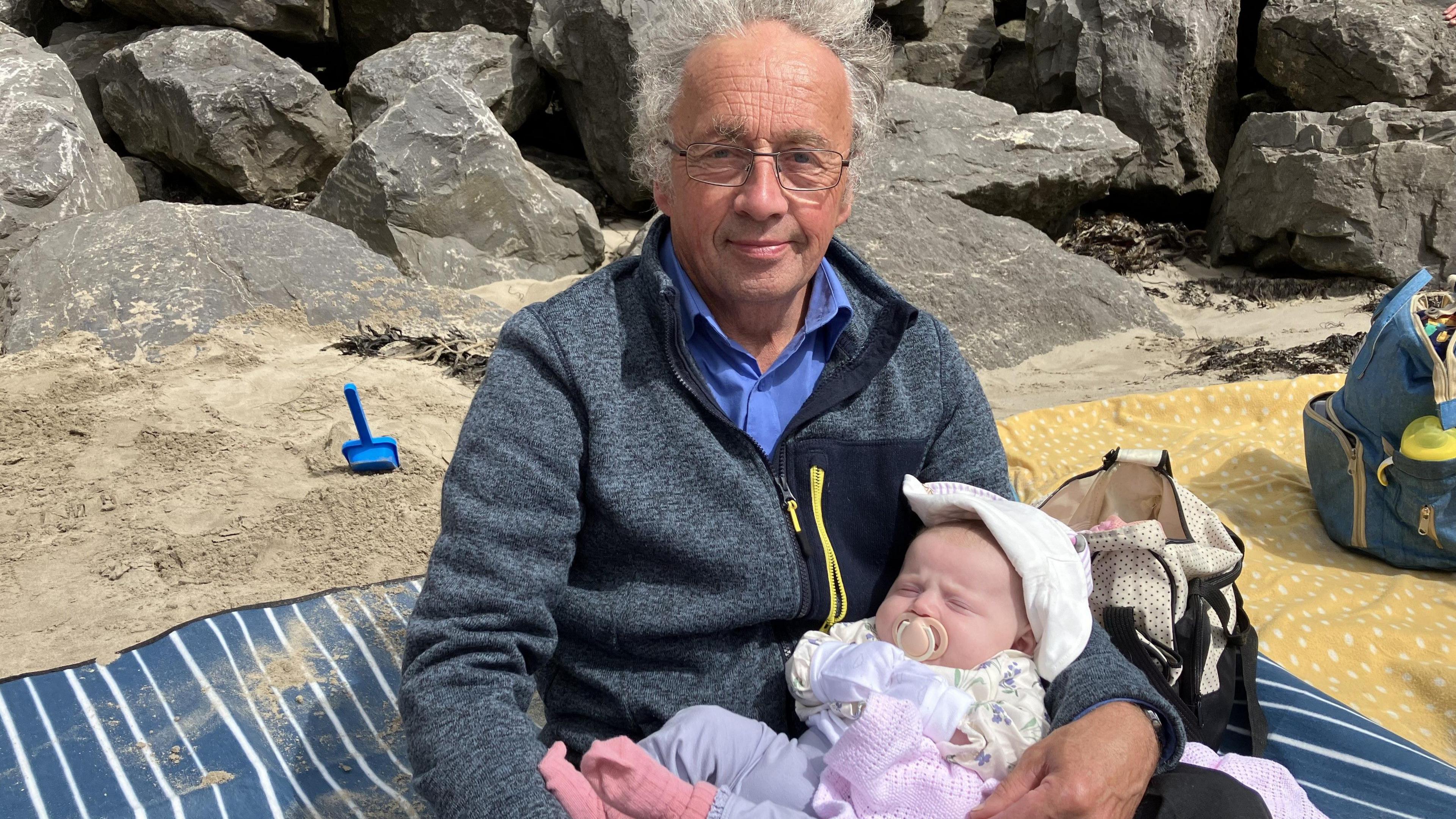 A man holds his sleeping granddaughter while they sit on a blanket at the beach