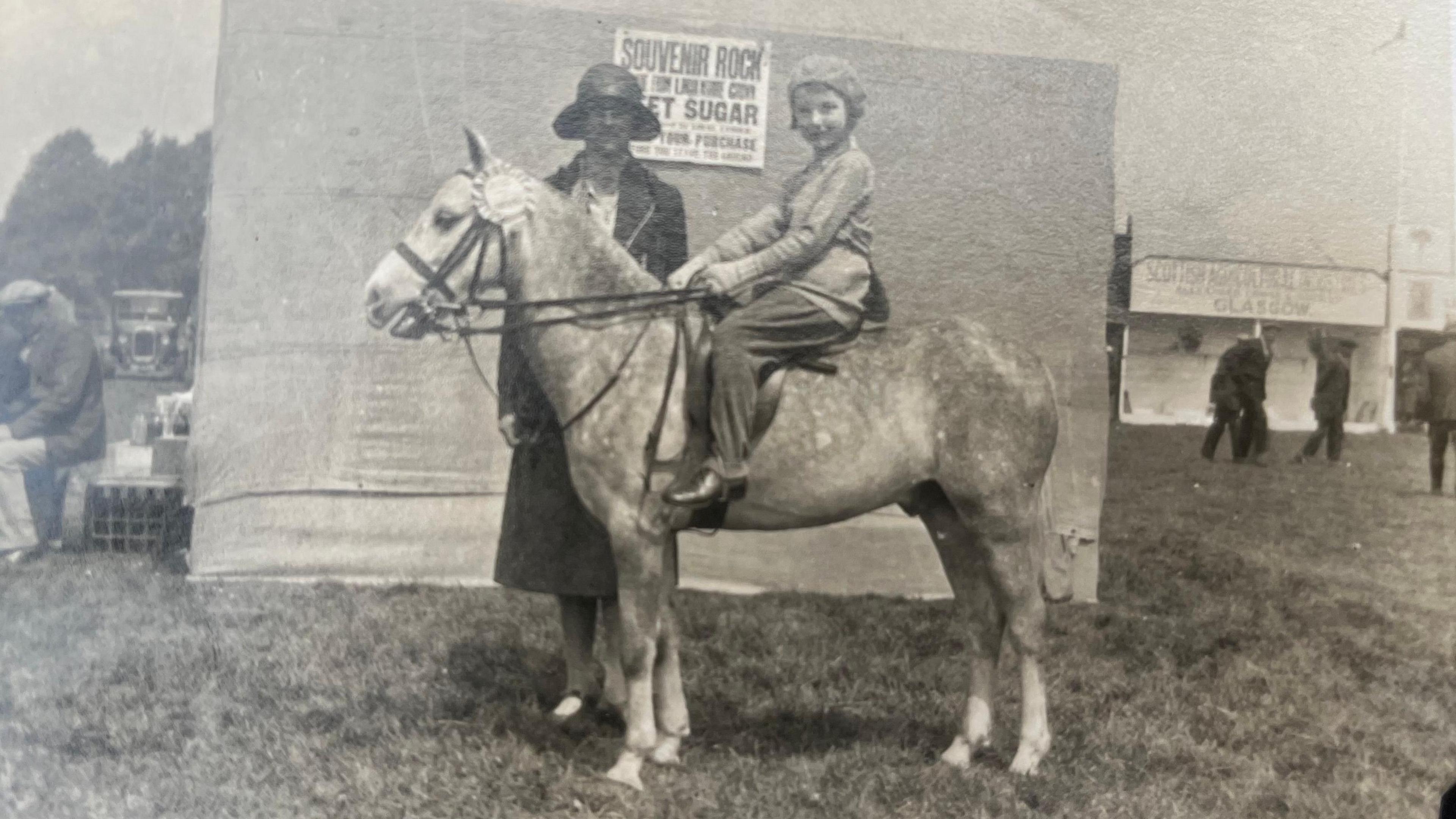 The image is a very old photo in sepia. A dappled grey pony is in the middle of the image and has a rosette on its bridle. A young girl is riding the pony and looking directly at the camera. She is wearing a beret style hat. Stood behind the pony is a women in a long dress and floppy hat, also looking at the camera. 