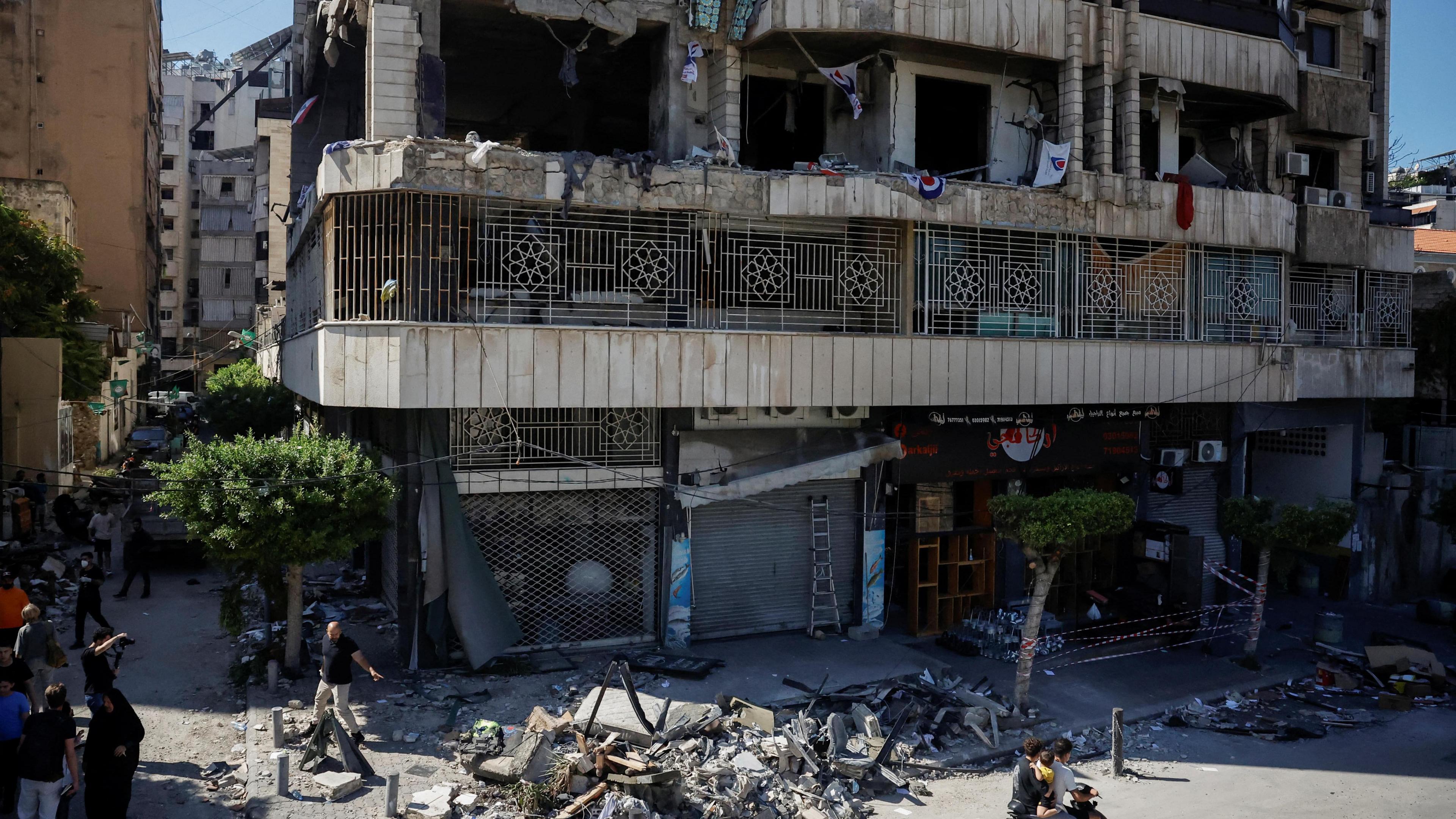 People walk past a concrete building with its windows blown out and rubble lying beneath.