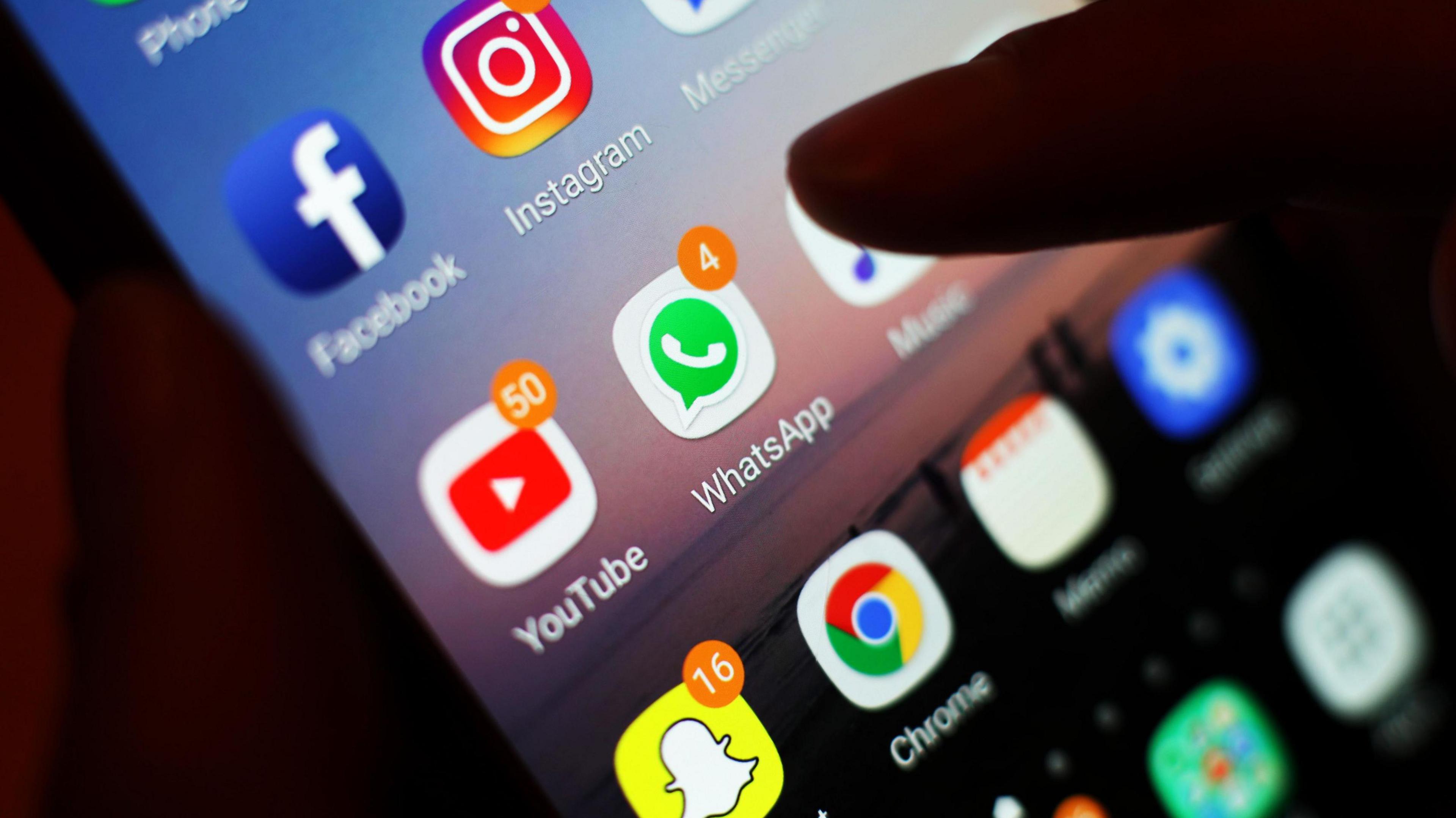 A mobile phone screen with the logos a range of internet services including the blue logo of facebook, yellow Snapchat logo and WhatsApp logo with a phone inside a green speech bubble. A finger is hovering over the link to music.