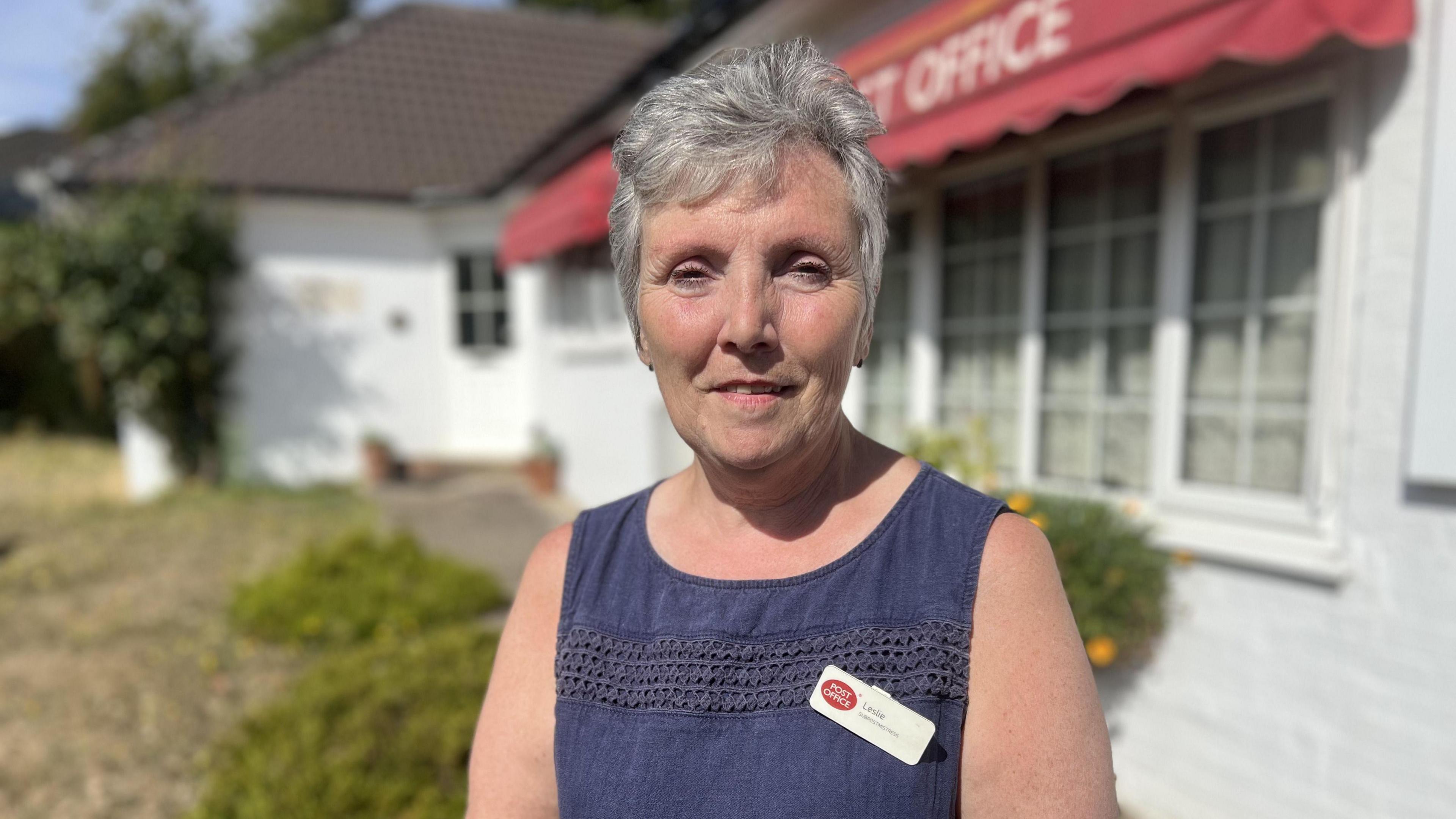 Leslie Barry has short grey hair and is wearing a blue top, with a post office name badge on. She is standing in front of a post office.