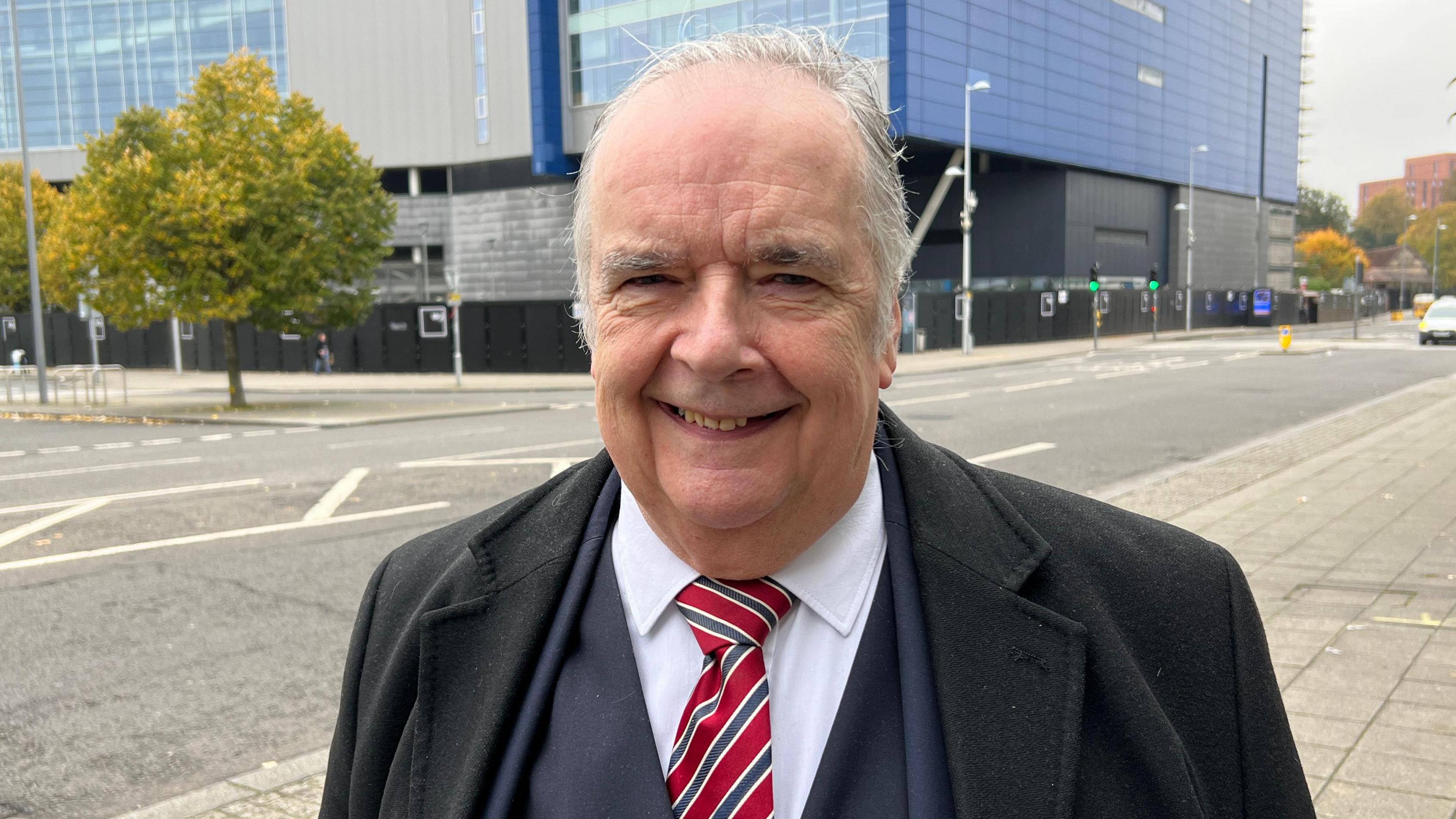 A man wearing a black overcoat on top of a navy suit with a red and grey striped tie looks towards the camera and smiles. He is standing in front of a road junction, a building and some trees are visible in the background