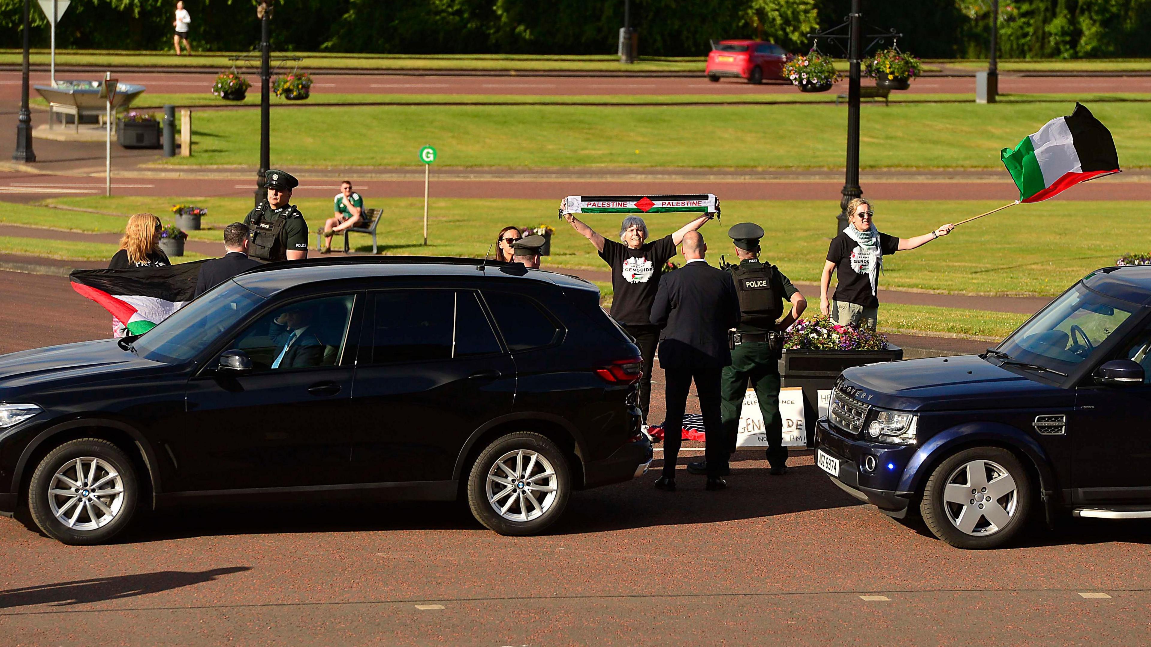 A gathering of people waving Palestine flags and holding up scarfs with the Palestinian colours as dark coloured vehicles drive by