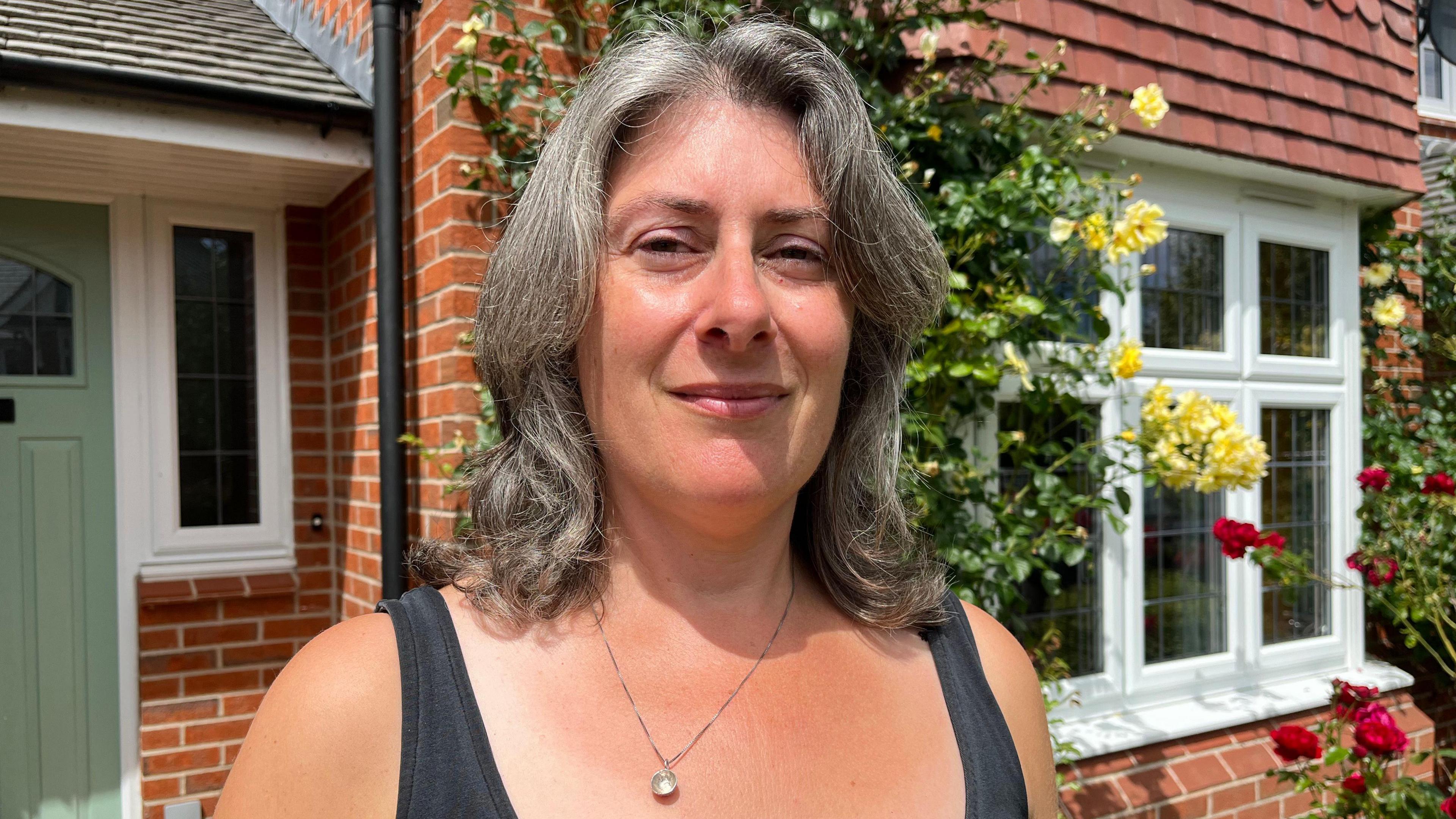 Jessica is standing in front of her house - a red-brick property with white windows, a sage green door, and red and yellow roses growing up the walls. She is wearing a black vest top, and is looking into the camera and smiling slightly.