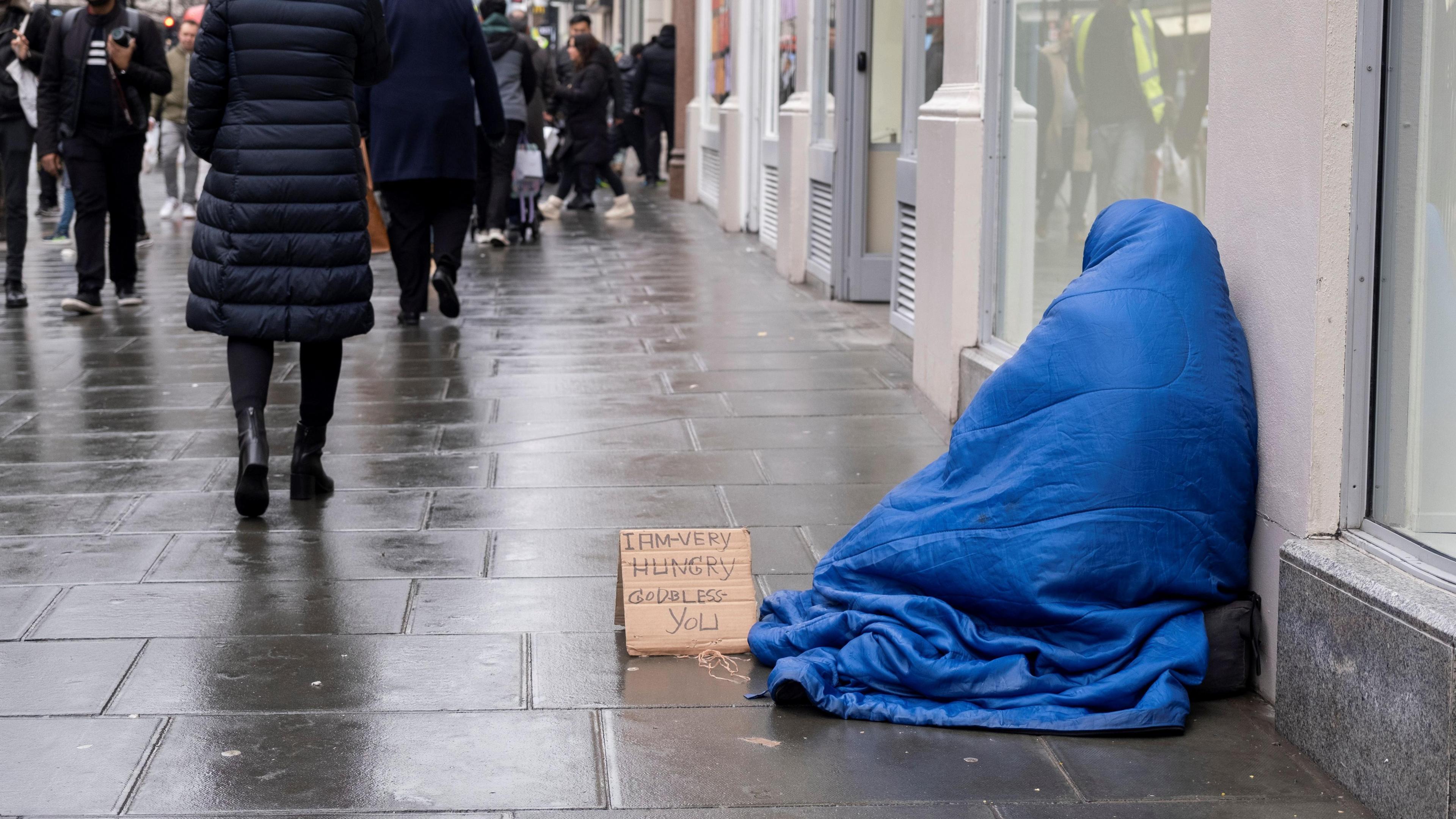 A street scene on a rainy day. A homeless person is on the right, sitting on the pavement and wrapped up completely in a blue sleeping bag. To his left is a small sign written on cardboard that says: "I am very hungry. God bless you." People can be seen walking along the street. 