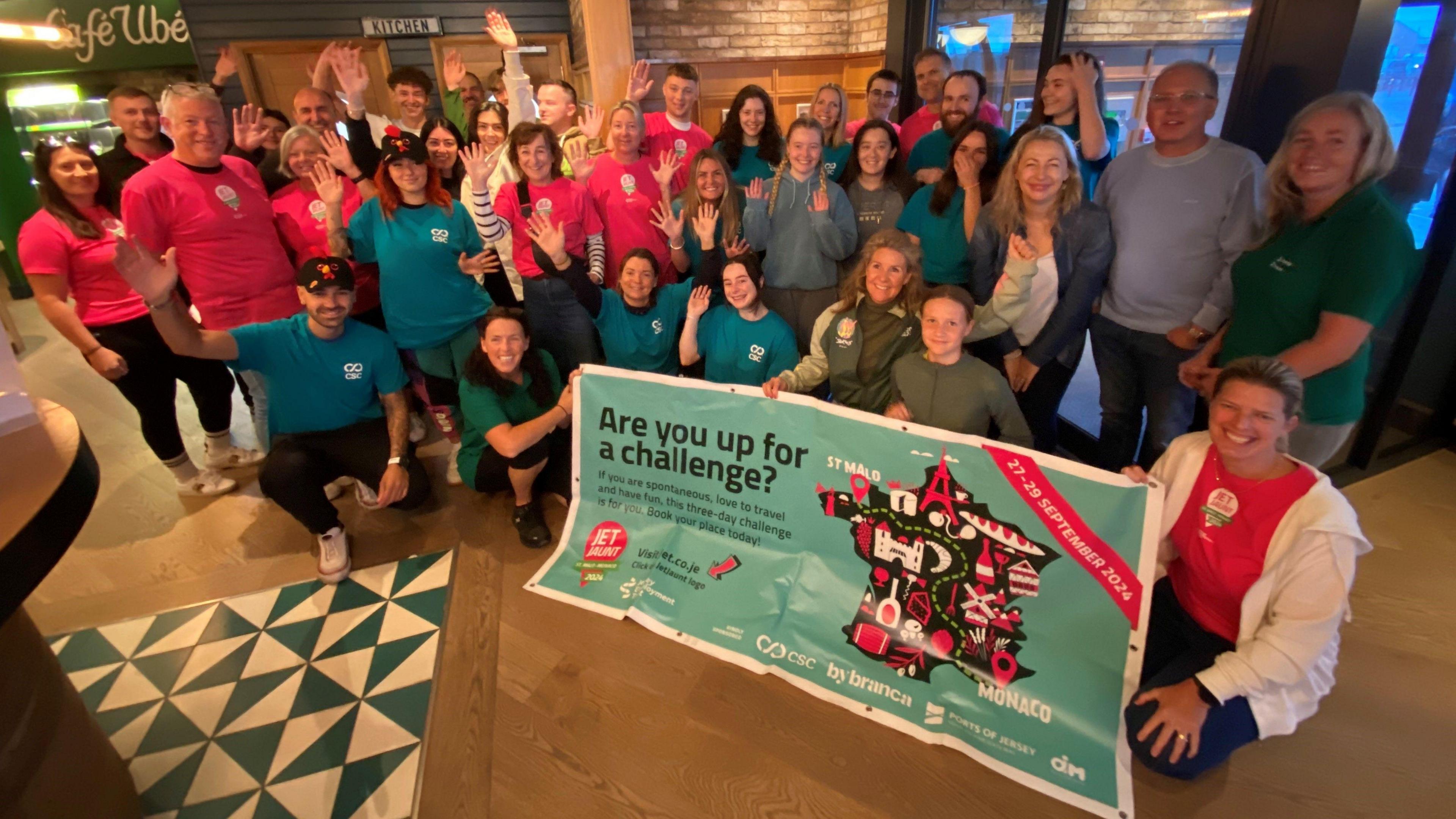 A group of people indoors pose together holding a large banner that reads 'Are you up for a challenge?' Some are wearing red shirts, others teal. The banner features logos for 'CoppaFeel!', 'boob or bust', 'MONDO', and 'dish', along with an illustration of a mountain with flags. In the background, there are signs labeled 'Kitchen' and 'Exit'.