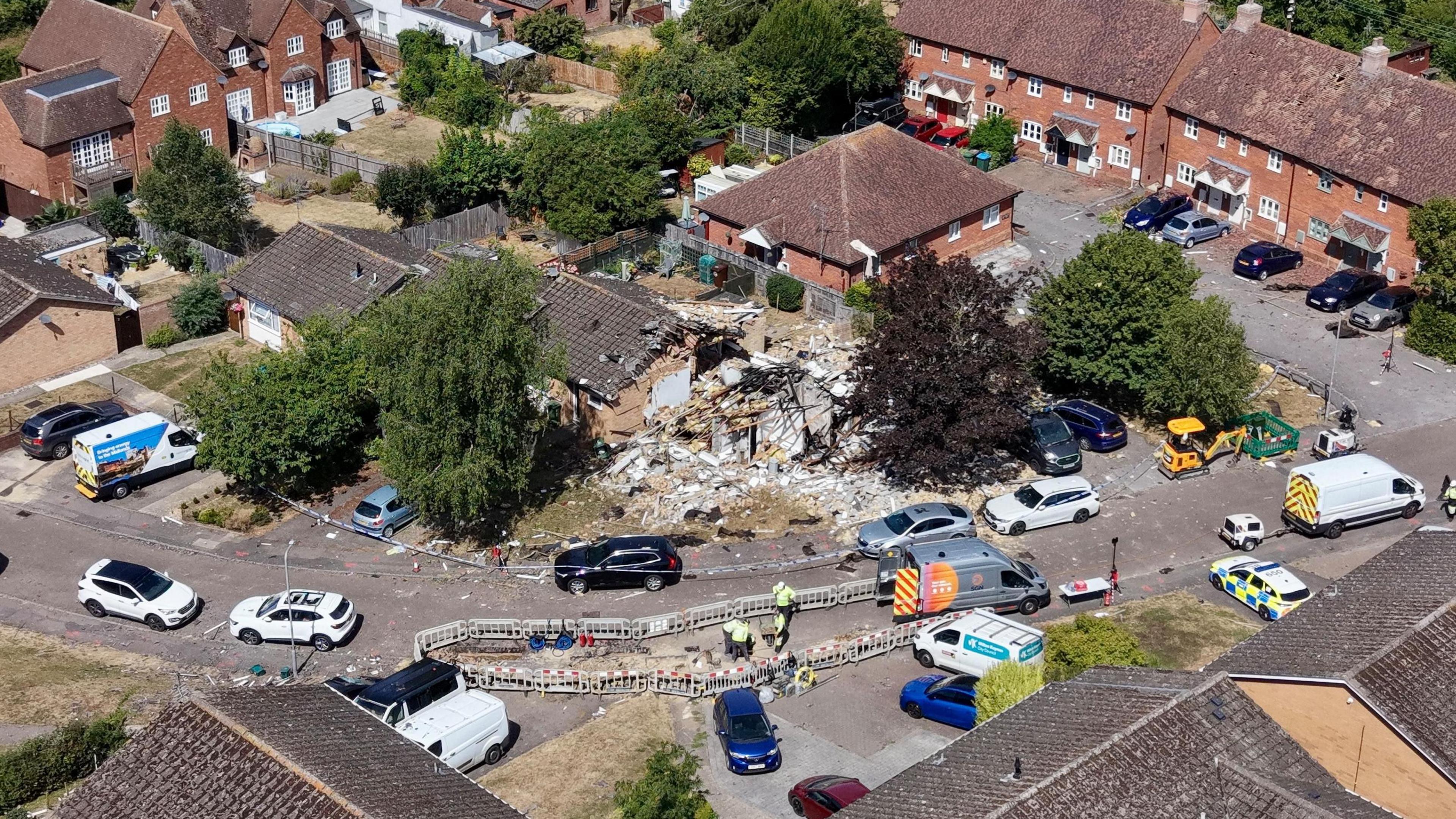 Aerial view of a house which has suffered an explosion and is completely destroyed. There is nothing but rubble and debris left. There are cars and vans parked on the road in the surrounding area. There are roadworks taking place on the road near the house.