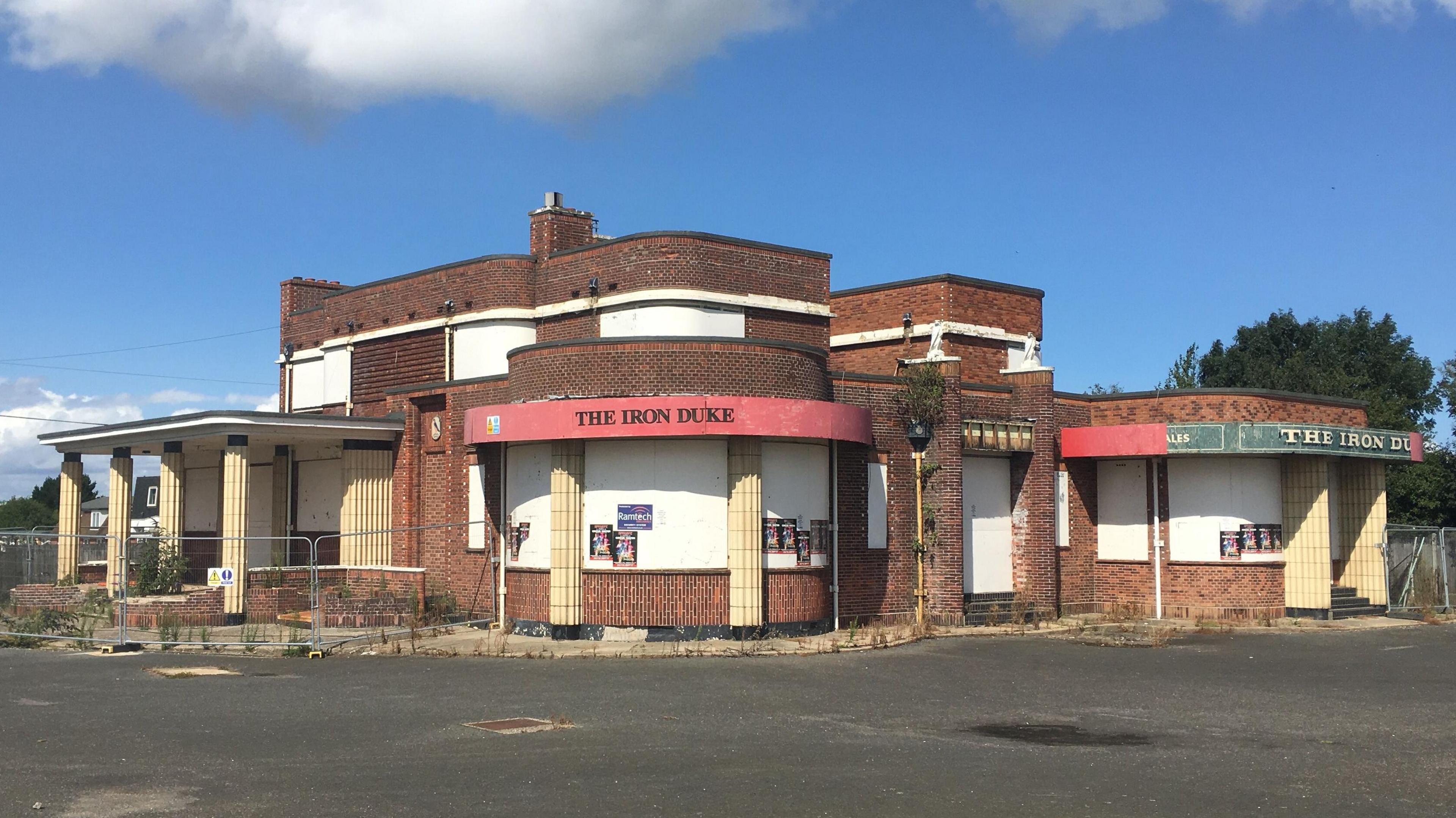 The Iron Duke, a former pub in Great Yarmouth. The building has been vacant and fencing is outside the building. Red signage on the pub says THE IRON DUKE in black writing.