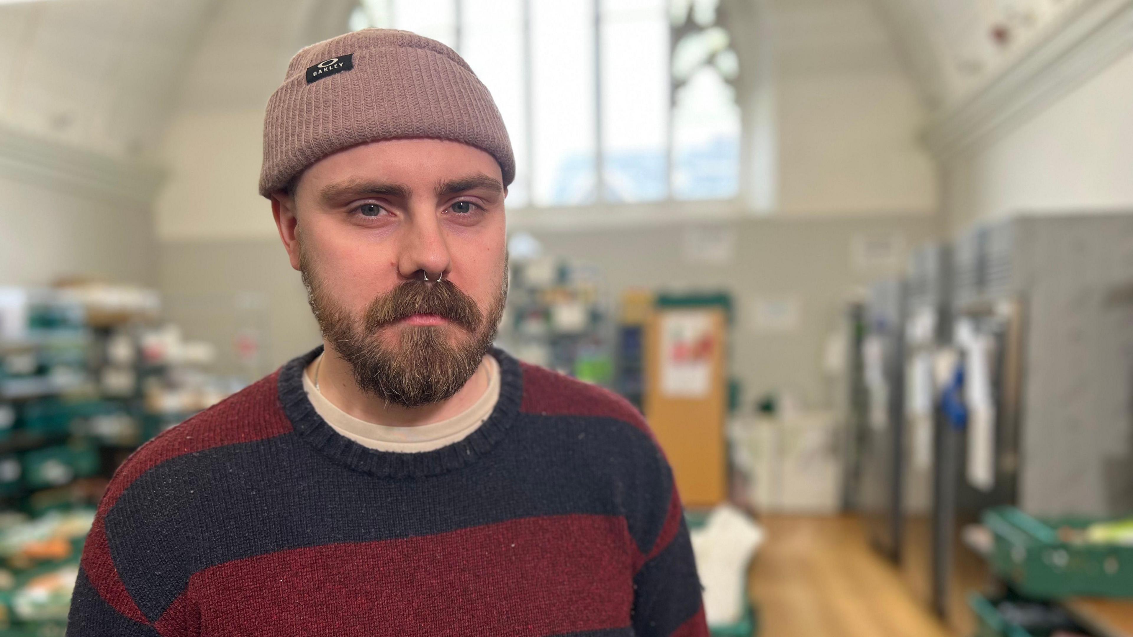 Daniel Cairney stands in a church building wearing a pink woolly hat and a red and black striped jumper.