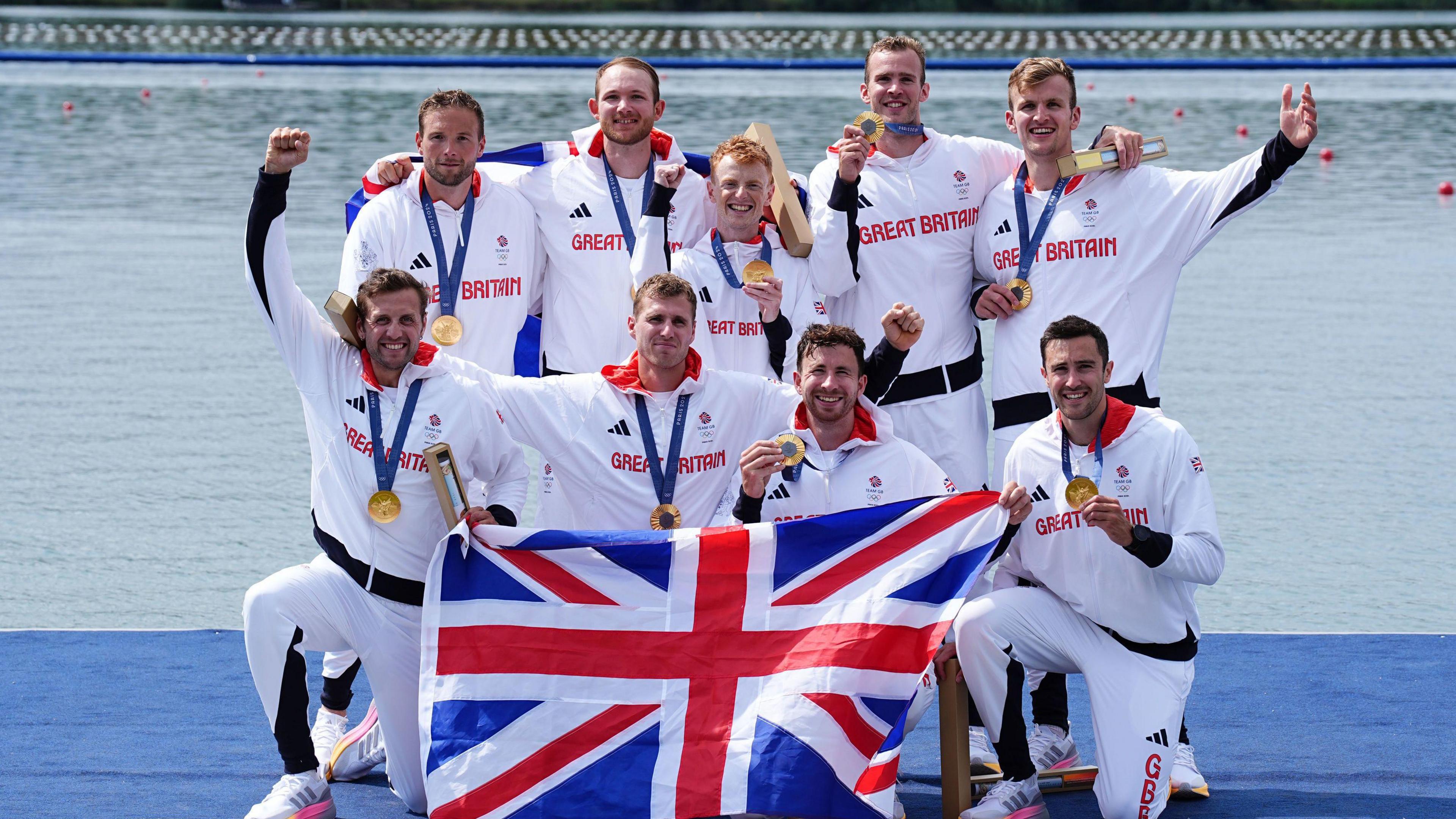 Eight men in white tracksuits with gold medals around their necks raise their arms in triumph holding up a union jack flag.