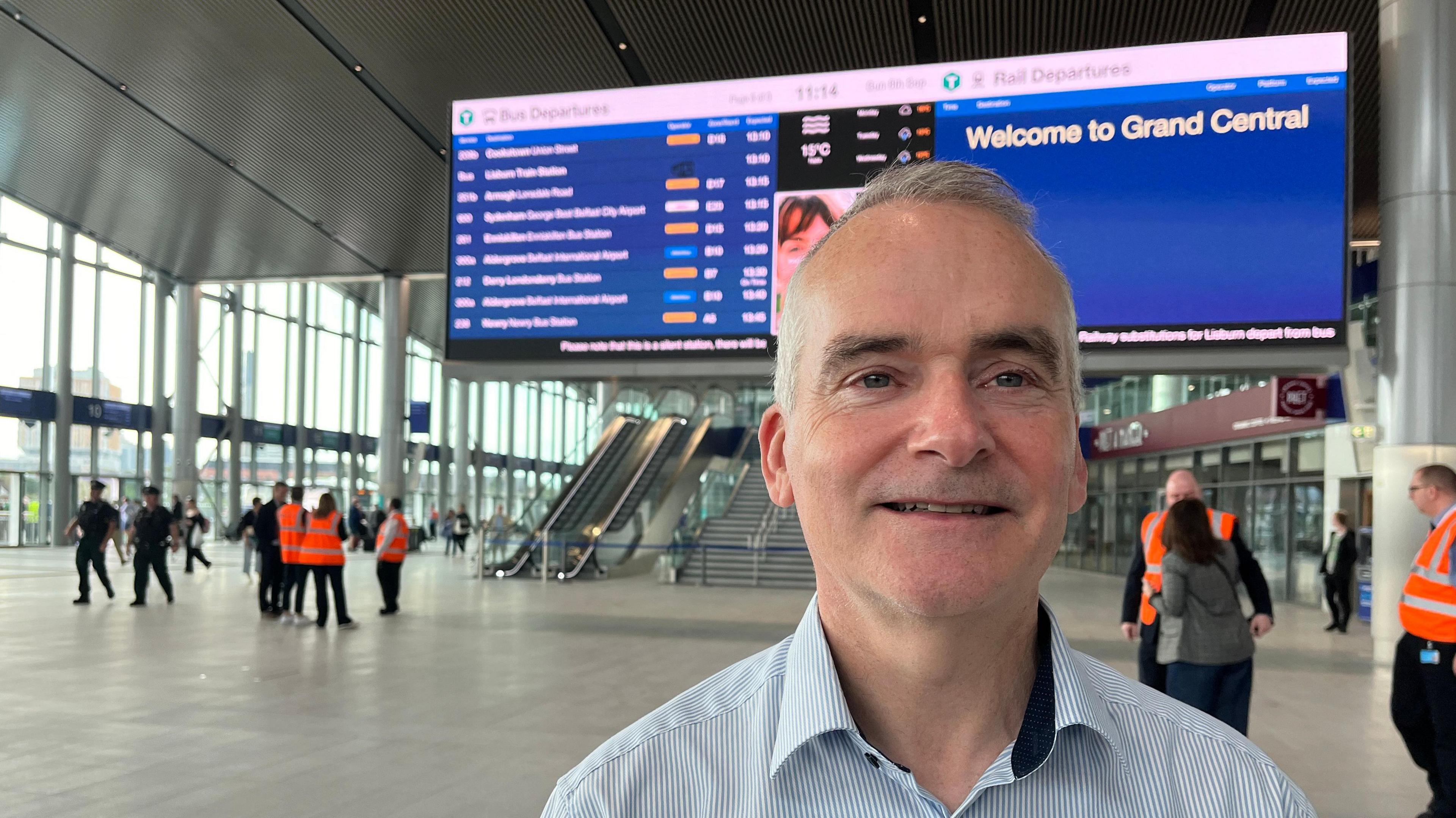 Translink Chief Executive Chris Conway standing in front of a passenger information sign in Belfast's Grand Central Station in September.  He has short, grey hair and is wearing a blue and white striped shirt with an open collar. 