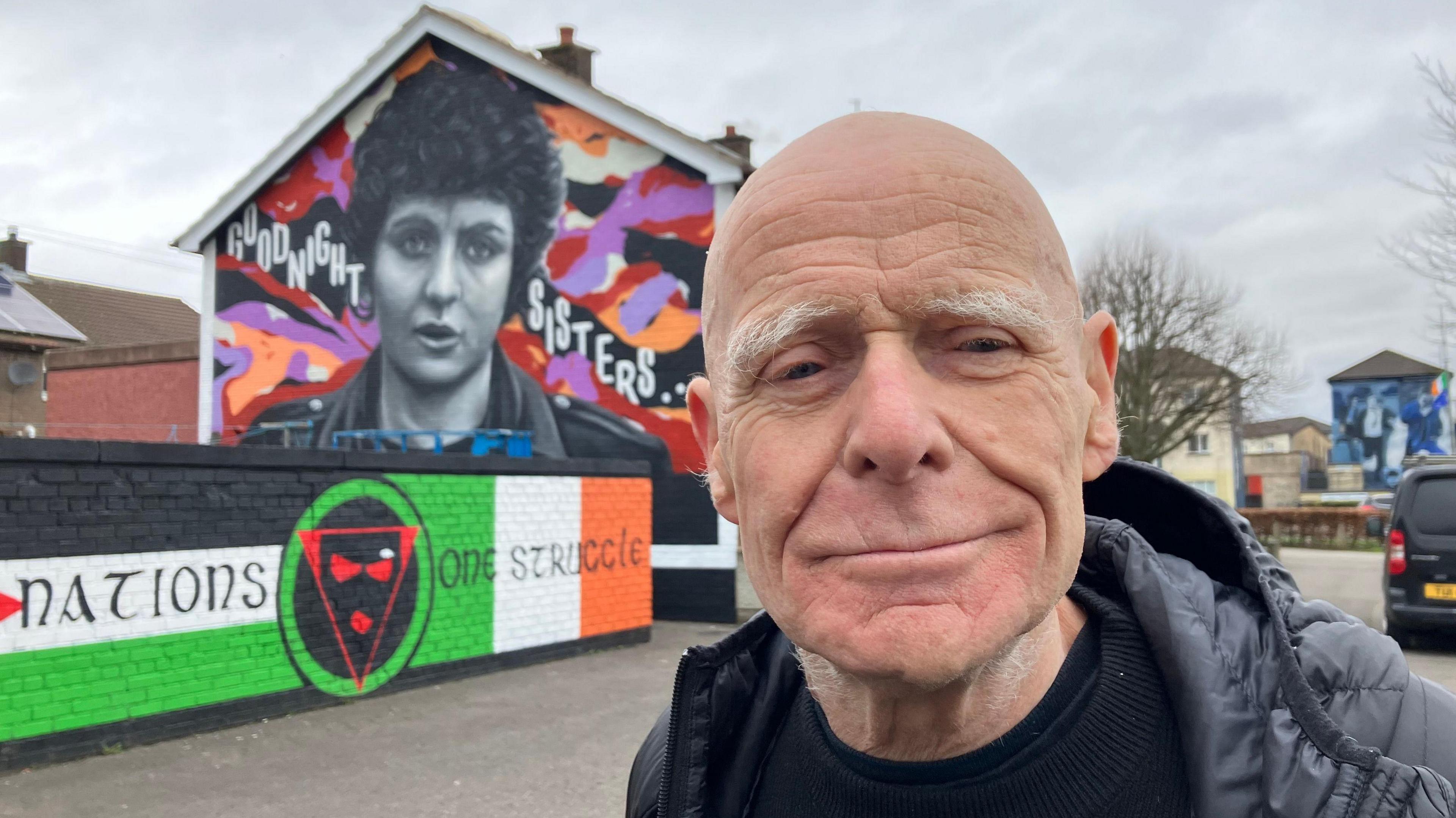 Eamonn McCann with the Nell McCafferty mural in the background. He is bald and wearing a black hooded padded jacket and black top. 