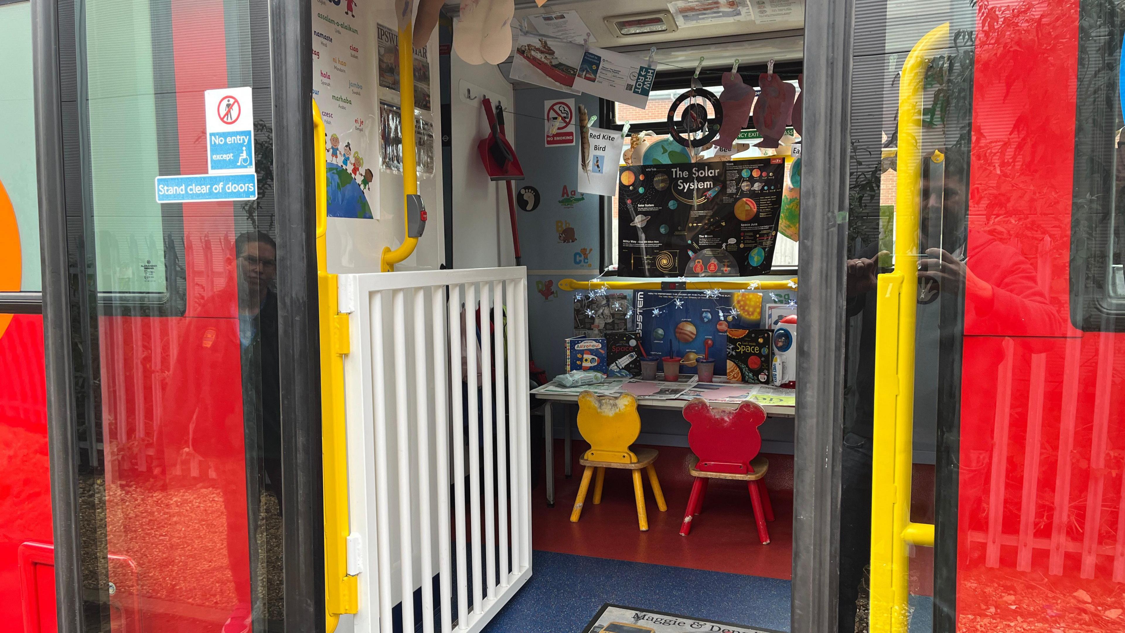 The view through open doors of a double decker bus which has been converted to have play equipment. Through the doors is a small desk with colourful chairs and space-themed posters.