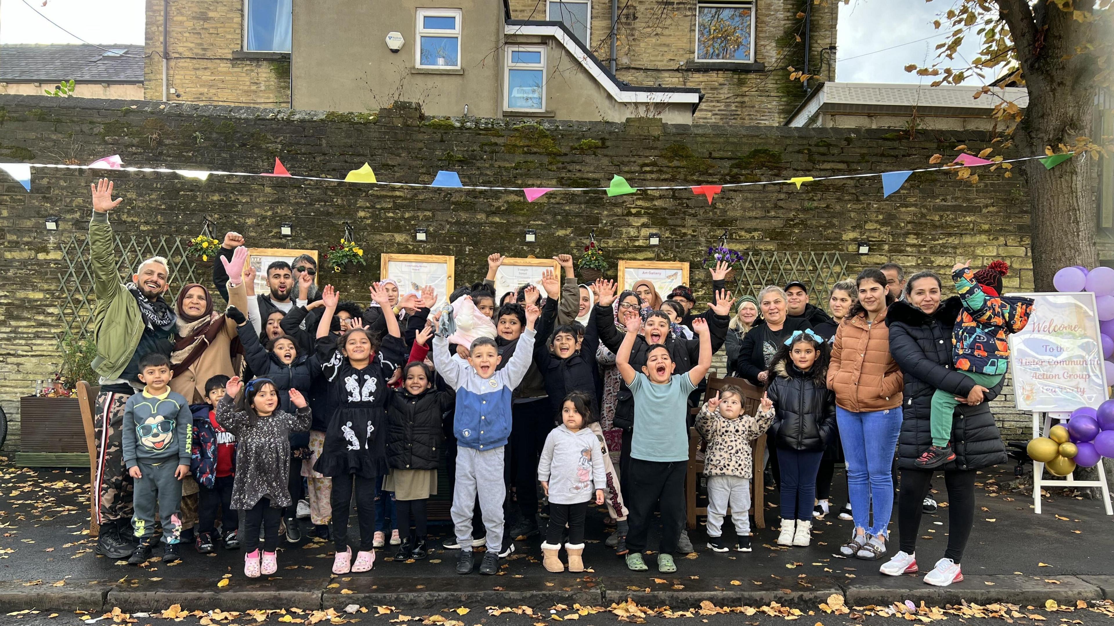 A large group of people including children posing in a celebration photograph in their street. There is bunting and balloons surrounding them.