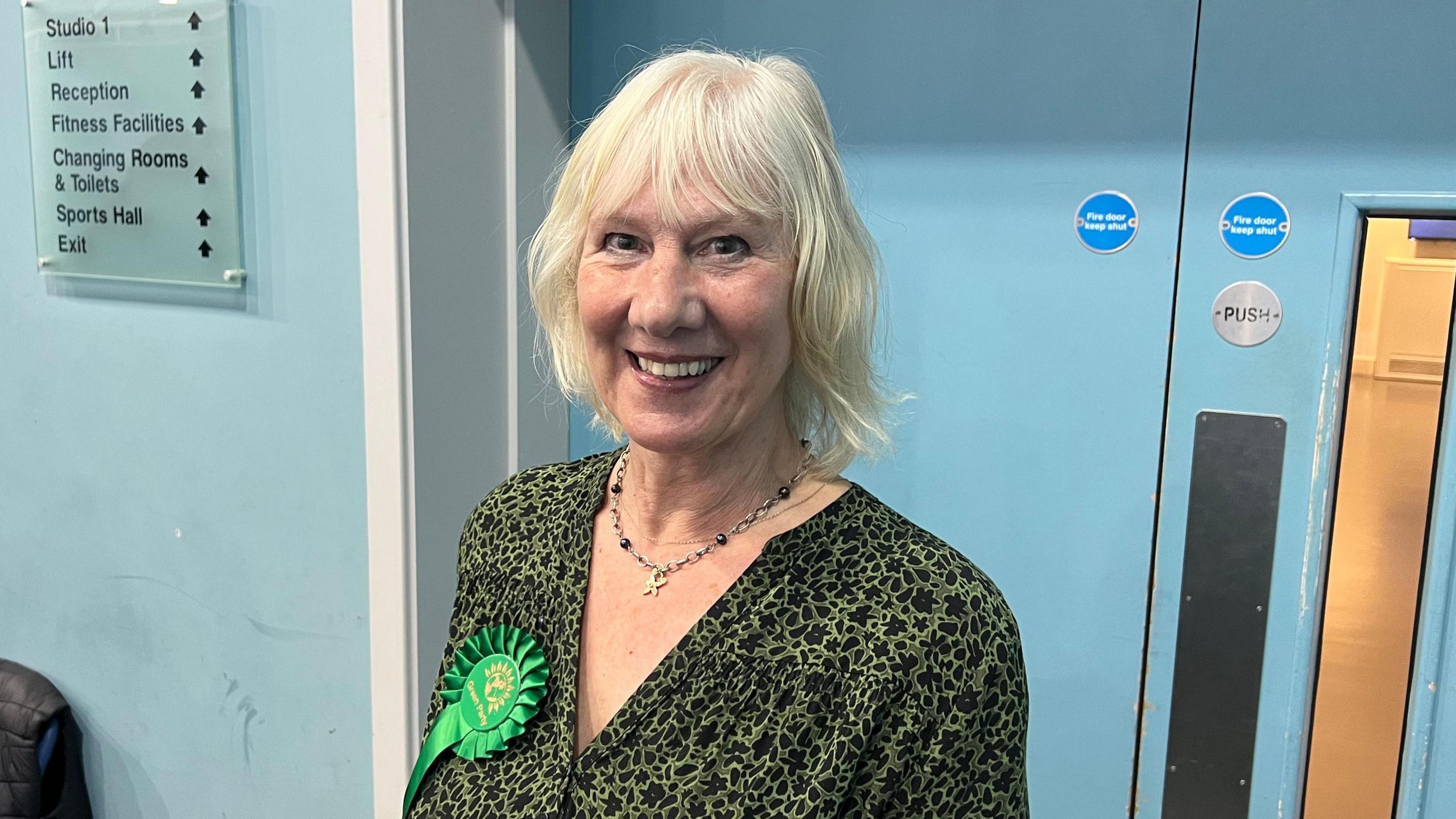 Jean Glasberg smiles at the camera as she is photographed standing in a corridor with blue walls. She has short blonde hair and is wearing a green and black patterned blouse with a green rosette on her right side. She is also wearing a necklace.